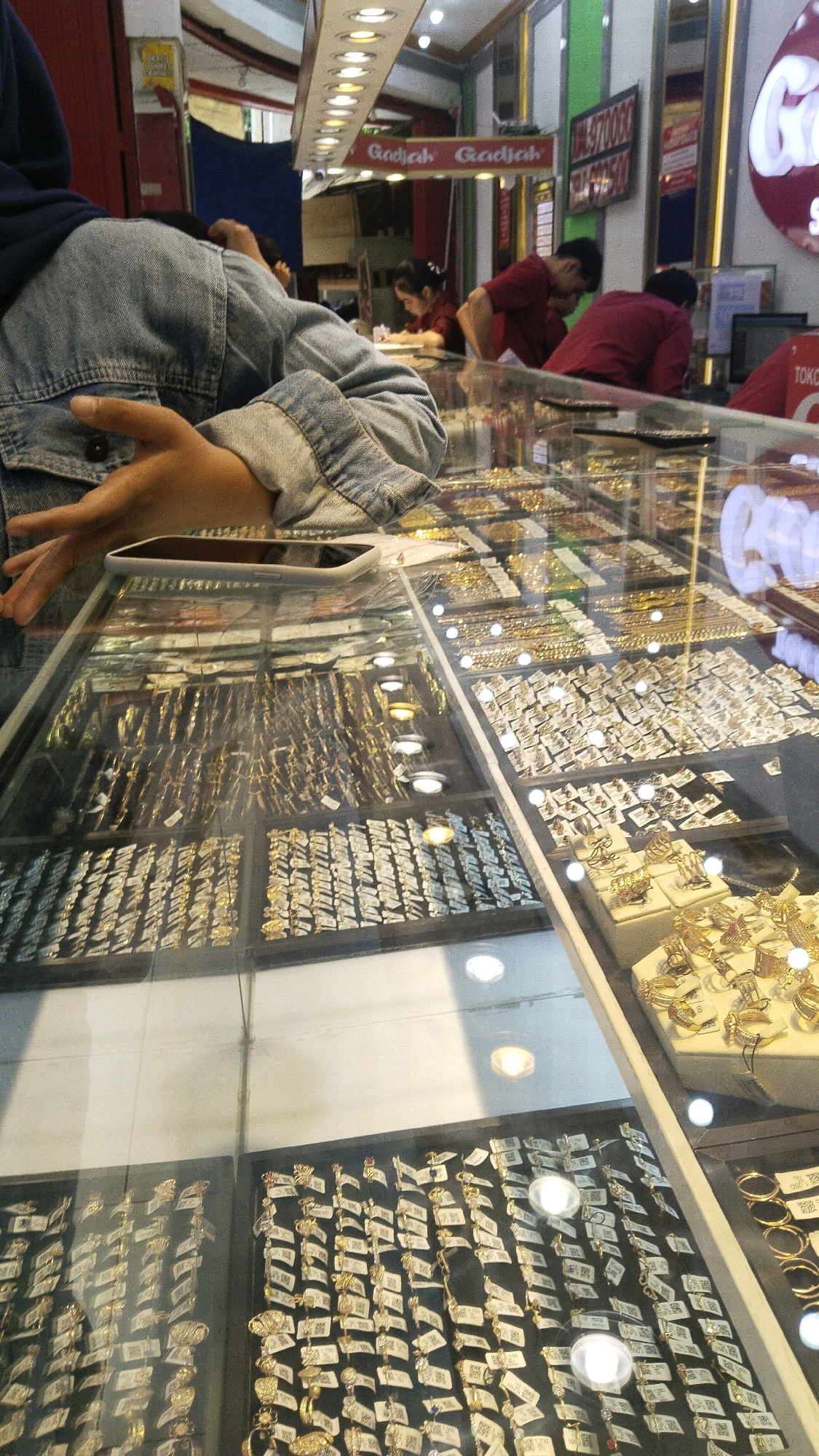 The image depicts the interior of a jewelry store in Jombang, Indonesia. The foreground is dominated by a glass display case filled with an extensive collection of gold jewelry, including rings, necklaces, and bracelets, neatly arranged and tagged. A person, partially visible from the shoulder down, is leaning over the display, their hand resting on the glass. In the background, several individuals are present. One person is wearing a dark blue headscarf and a denim jacket, and another is wearing a red shirt. Other people in red shirts are seen interacting behind the counter. The store appears well-lit with overhead lighting. A sign with the text "Gajah Gajah" is visible on the wall, suggesting the store's name. The overall scene conveys a busy commercial environment focused on the sale of gold jewelry.