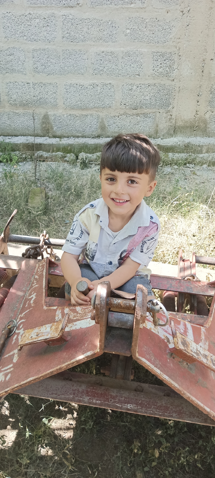 A young boy is sitting on a piece of rusty farm equipment in an outdoor setting. The boy has dark hair with bangs and is wearing a collared shirt. He is smiling and looking directly at the camera. The farm equipment is red and rusty, with various metal parts and chains. Behind the boy, there is a wall made of grey concrete blocks. The ground is covered with grass and weeds. The location is the city of Unknown, Unknown.