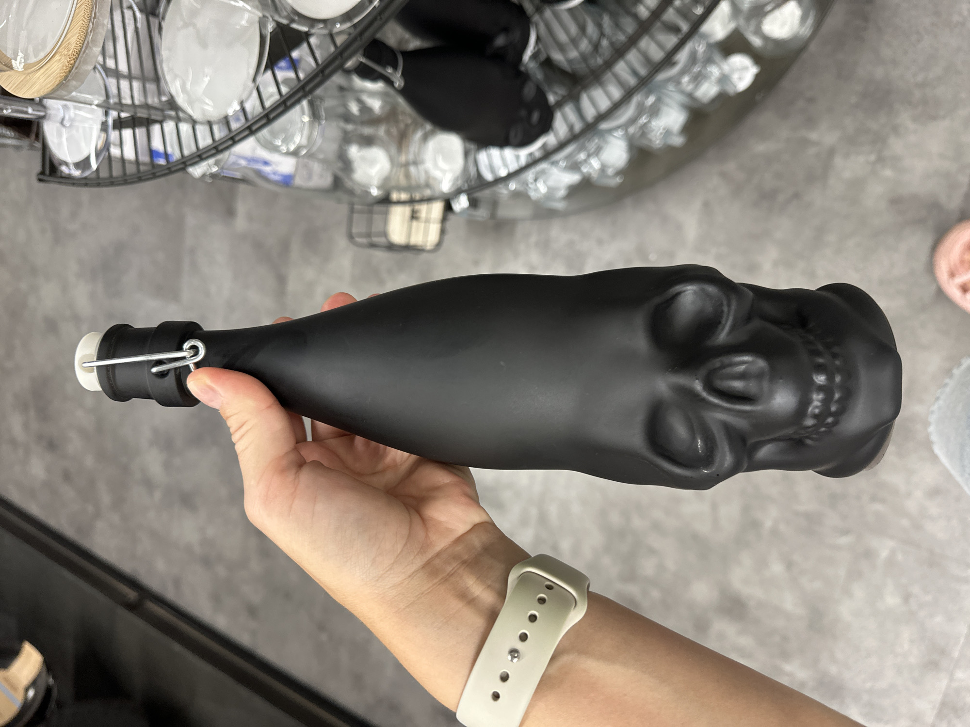 A person holds a black skull-shaped bottle with a stopper in their right hand, wearing a light-colored smartwatch. The background shows a display of kitchenware, including glasses and storage containers, in a retail setting. The location is likely a home goods store in Stade, Germany.