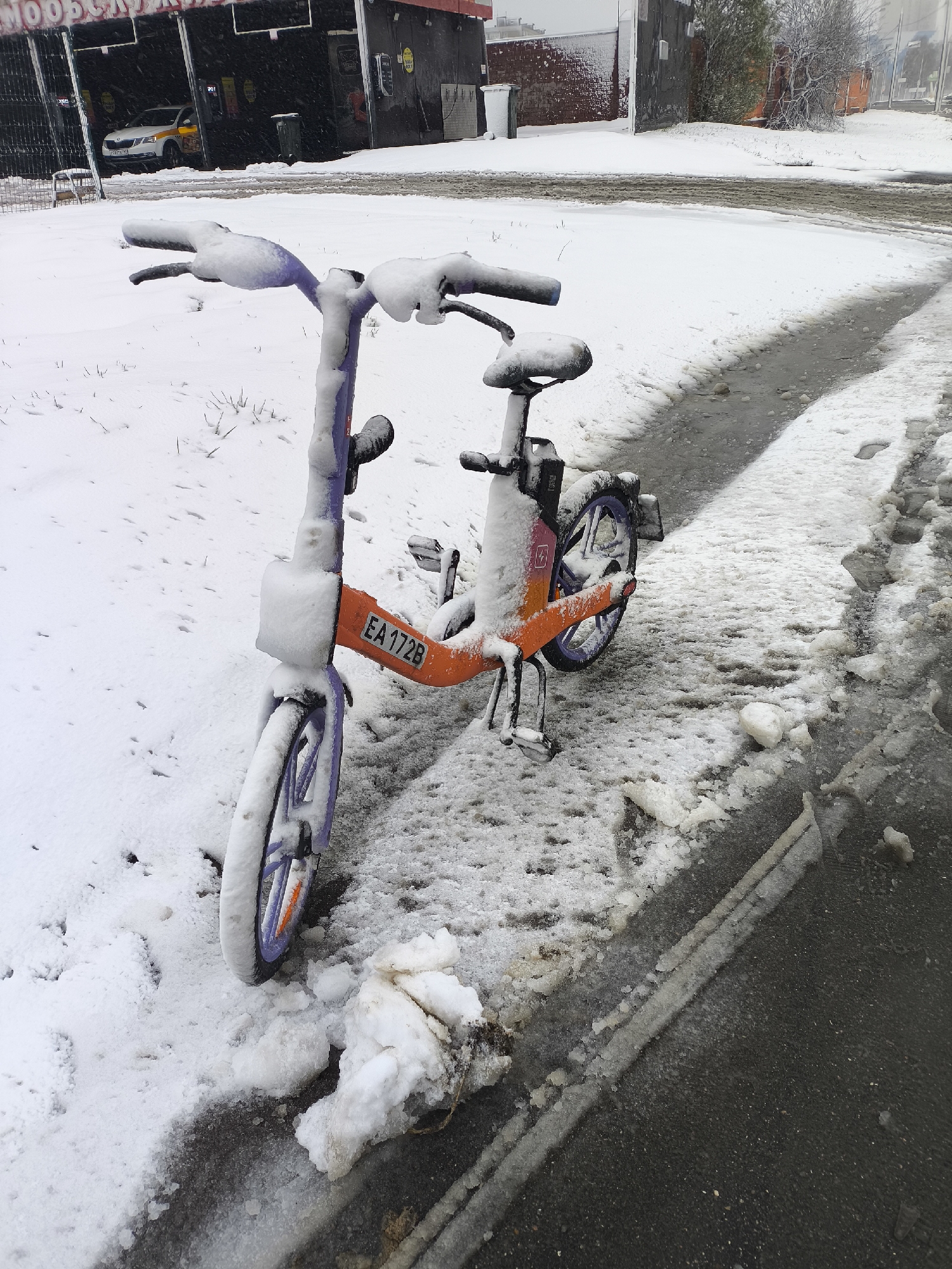 An outdoor winter scene captures a bright orange and purple shared bicycle, largely covered in fresh snow, parked on a snow-covered verge adjacent to a partially cleared asphalt path. The bike's front stem displays the identifier "EA 172B," and a lightning bolt symbol within a square is visible on its rear frame. The ground is a mix of untouched snow, slush, and icy patches, indicating recent and ongoing snowfall. In the background, a dark building, likely a self-service car wash, features the partial text "МОЙОСЛУЖБИ" (likely part of "МОЙКА САМООБСЛУЖИВАНИЯ" - self-service car wash in Russian) on its facade. A white car, possibly a taxi, is visible inside one of the open bays. The sky is overcast, suggesting daytime with heavy cloud cover. The scene depicts a cold, snowy day in an urban environment, consistent with winter conditions in Moscow, Russia. No people are visible.
