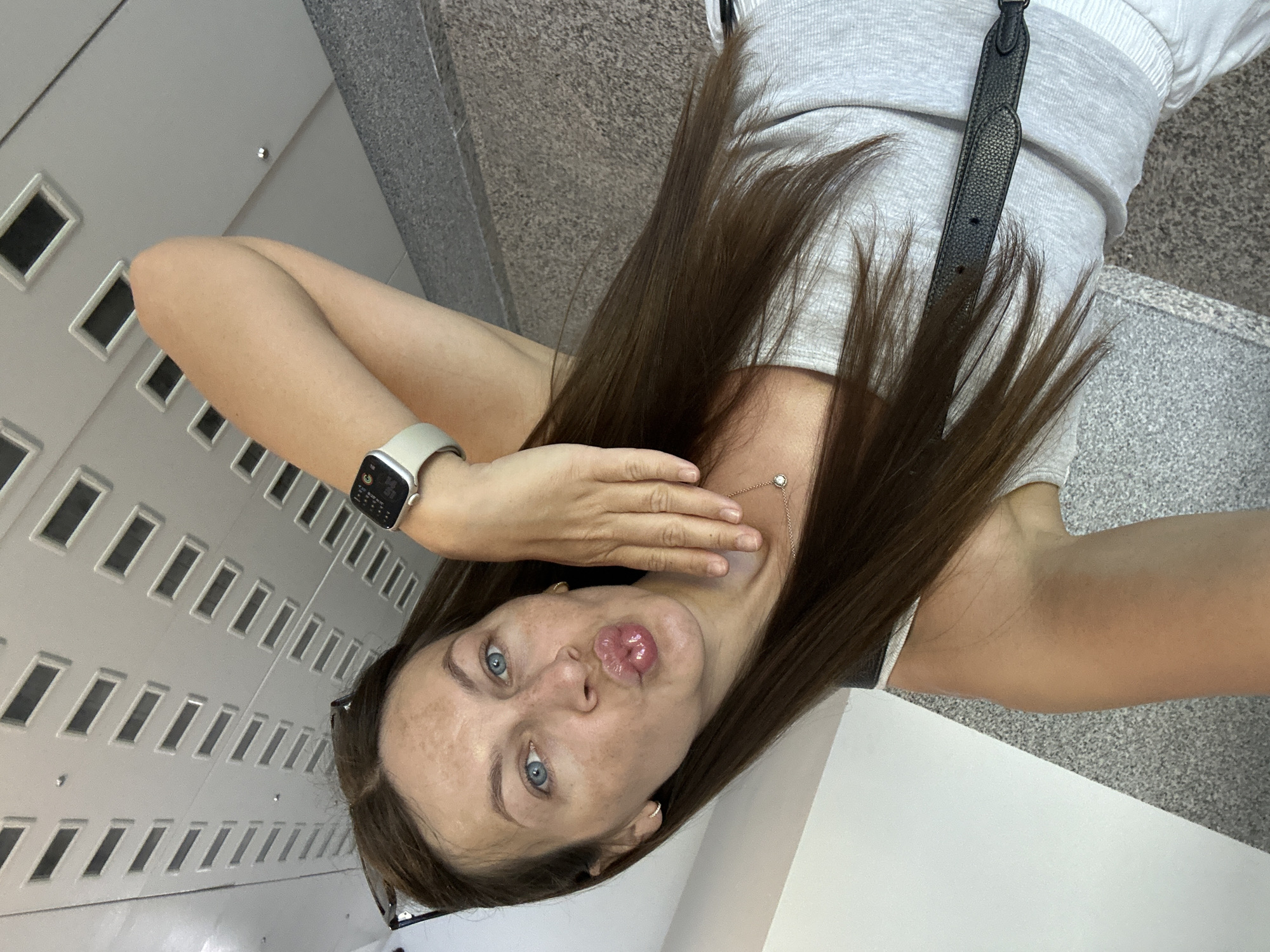 A woman with long brown hair and blue eyes is posing for a selfie. She has fair skin with freckles and is wearing a smartwatch on her left wrist. Her lips are pursed in a kissing gesture. She is wearing a gray top with a black strap. The background appears to be an interior space with a wall featuring a pattern of rectangular openings. The location is not evident in the image.