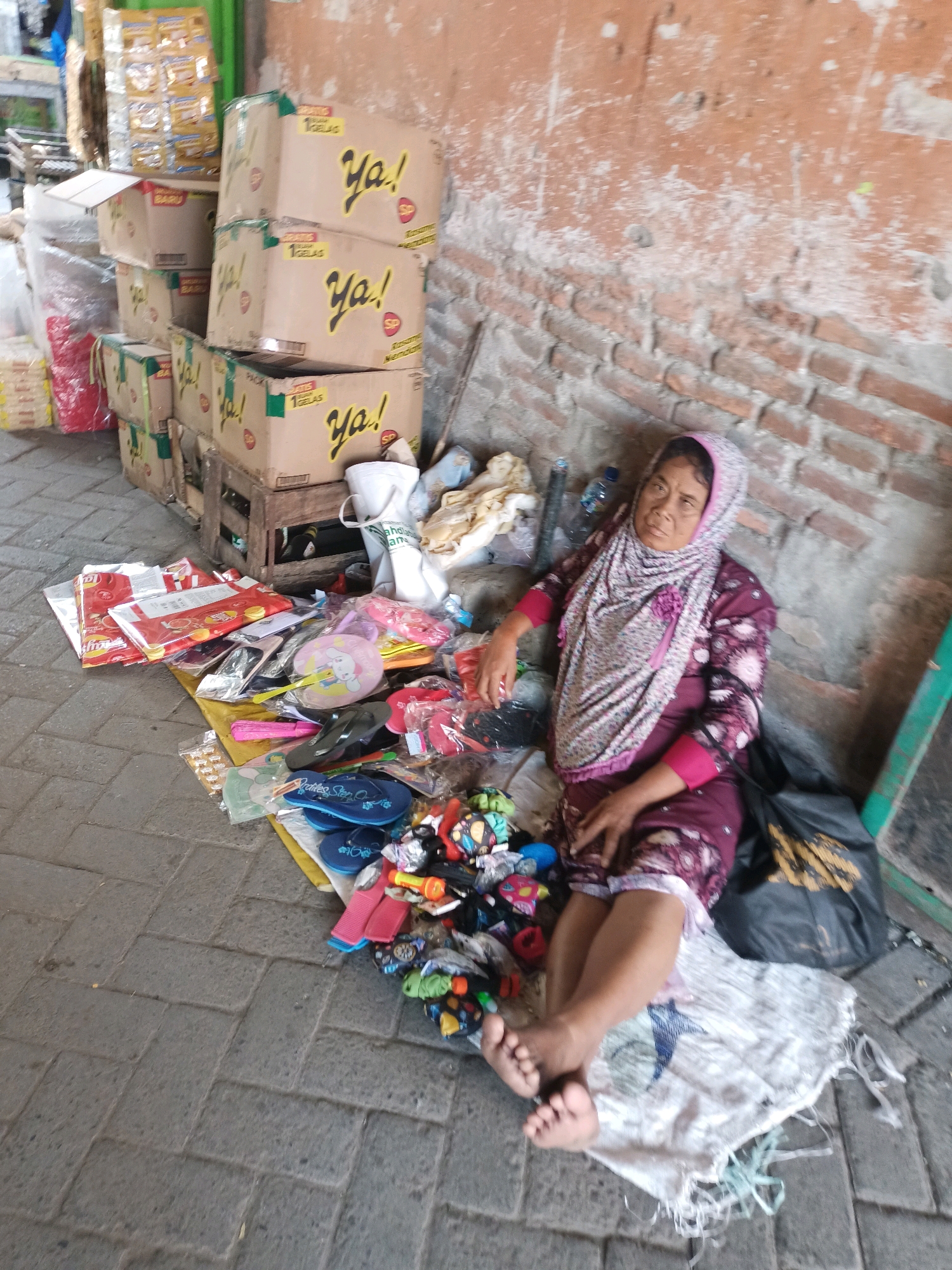 In Badas, Indonesia, a woman wearing a headscarf and a maroon patterned dress is seated on a rough, soiled white cloth. She is positioned against a brick wall with a weathered, orange-red plaster exterior, and her legs are extended. Beside her, a black plastic bag with a yellow pattern rests on the ground. The scene is cluttered with various items. Piles of cardboard boxes, some bearing the text "Ya!", are stacked to her left. Scattered on the ground in front of her are numerous small objects, including several pairs of flip-flops, small toys, hair accessories, and packaged snacks. A few plastic bags and loose clothing are also visible amongst the debris. The ground is paved with gray interlocking bricks.