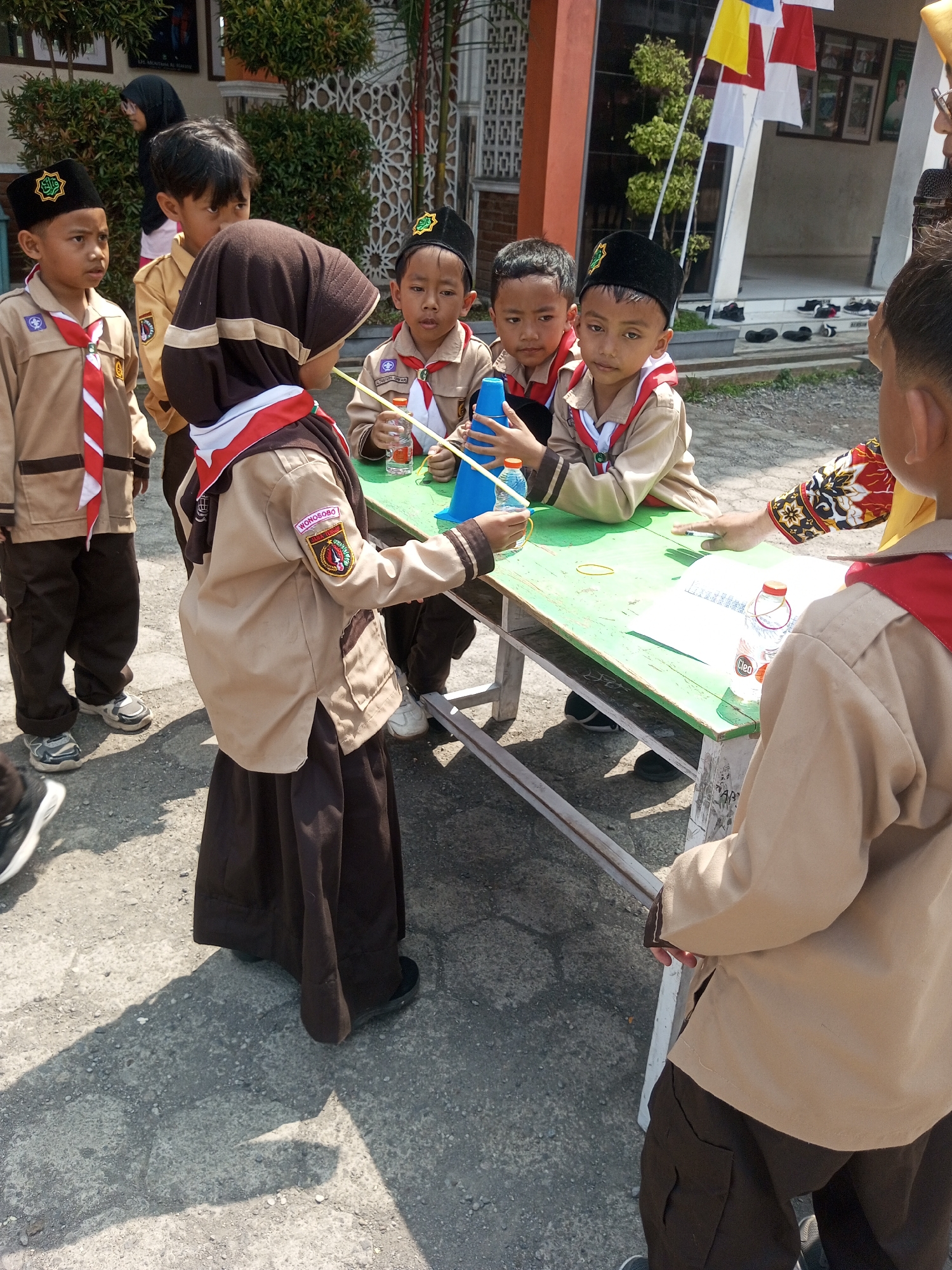 The image depicts an outdoor scene in the city of Kalibeber, Indonesia, possibly near a school, as suggested by the children's uniforms. Several young students, dressed in tan uniforms with red and white neckerchiefs and black caps, are gathered around a green table. One student, wearing a brown headscarf and a tan uniform, is holding a blue conical object. Another student to their left is wearing a black cap with a green emblem and a tan uniform. To the right, a student in a similar uniform holds a yellow stick, and a partially visible adult, also in uniform, appears to be interacting with the children. On the table, there are papers, a bottle of water with a red cap, and rubber bands. The ground is paved with gravel. In the background, there is a building with orange and white elements, and foliage. Several small flags, red, white, and yellow, are visible hanging in the background. The lighting suggests a sunny day, with distinct shadows on the ground.