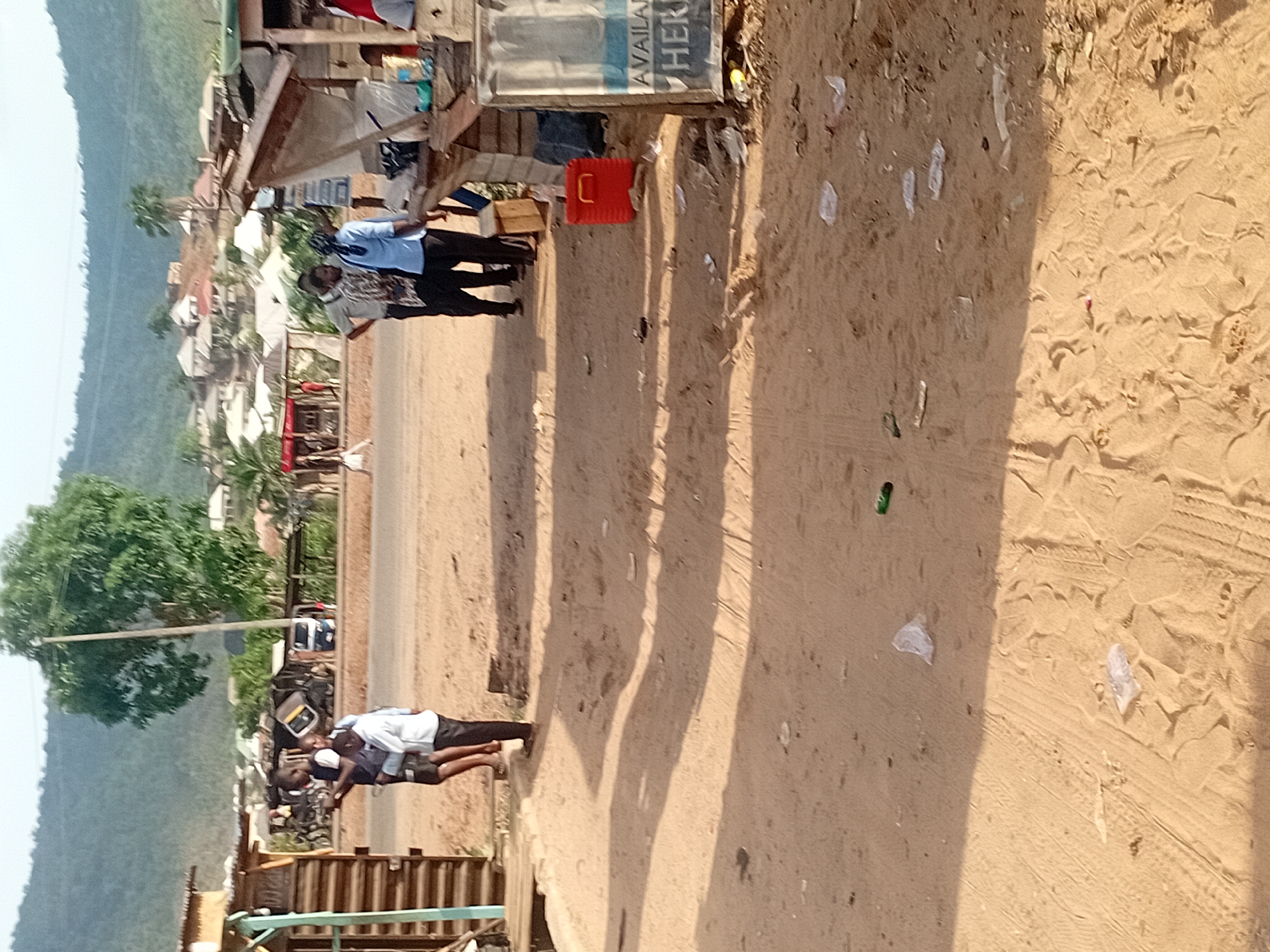 This photograph depicts a lively street scene, likely in Lokoja, Nigeria, during what appears to be a bright, sunny day. The clear sky and distinct, elongated shadows cast across the ground suggest it is either morning or late afternoon.
In the immediate foreground, a wide, sandy path is visible, marked by various footprints and tire tracks. The path is littered with scattered debris, including several white plastic wrappers and a green glass bottle.
Mid-ground, running horizontally across the frame, is a dirt road or wider pathway. Adjacent to this path, on the left, are several makeshift structures constructed from wood and possibly corrugated iron, resembling market stalls or small roadside shops. Near one of these structures, a man in a light blue collared shirt and dark trousers stands, appearing to interact with another individual. A red plastic crate is also visible on the ground beside the structure. Further down the path, another group of people can be seen near similar structures.
Beyond these foreground elements, a paved road is discernible, followed by more permanent buildings and abundant green vegetation. In the background, lush green hills or mountains rise majestically against the horizon. A large, verdant tree stands prominently on the left side of the scene.
A partial sign on one of the makeshift structures displays the text "AVAILA" and "HER", likely part of "AVAILABLE HERE". The overall scene portrays a bustling, informal commercial or residential area.