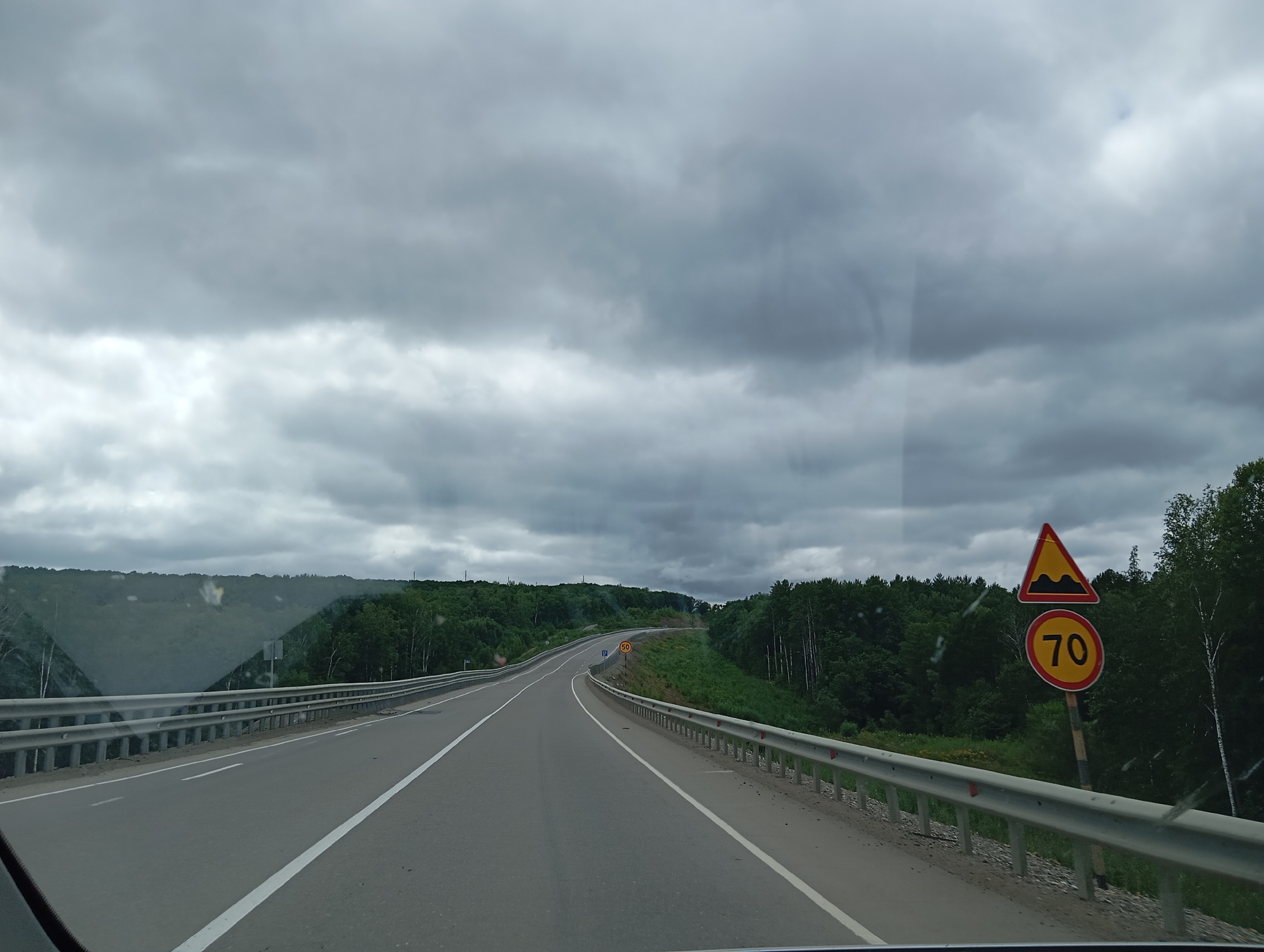 The image depicts a two-lane asphalt road curving through a densely forested, hilly landscape under a heavily overcast sky. A metal guardrail runs along the right side of the road. A yellow triangular sign with a red border depicts a series of bumps, indicating a bumpy road. Below it, a circular sign with a red border displays the number "70" in black, signifying a speed limit of 70 kilometers per hour. The environment suggests a rural or semi-rural setting, possibly in or near Taezhnoe, Russia, given the dense coniferous and deciduous forests characteristic of the Siberian taiga. There are no visible people or vehicles other than the perspective of being inside a moving vehicle. The overall atmosphere is muted due to the cloudy weather.