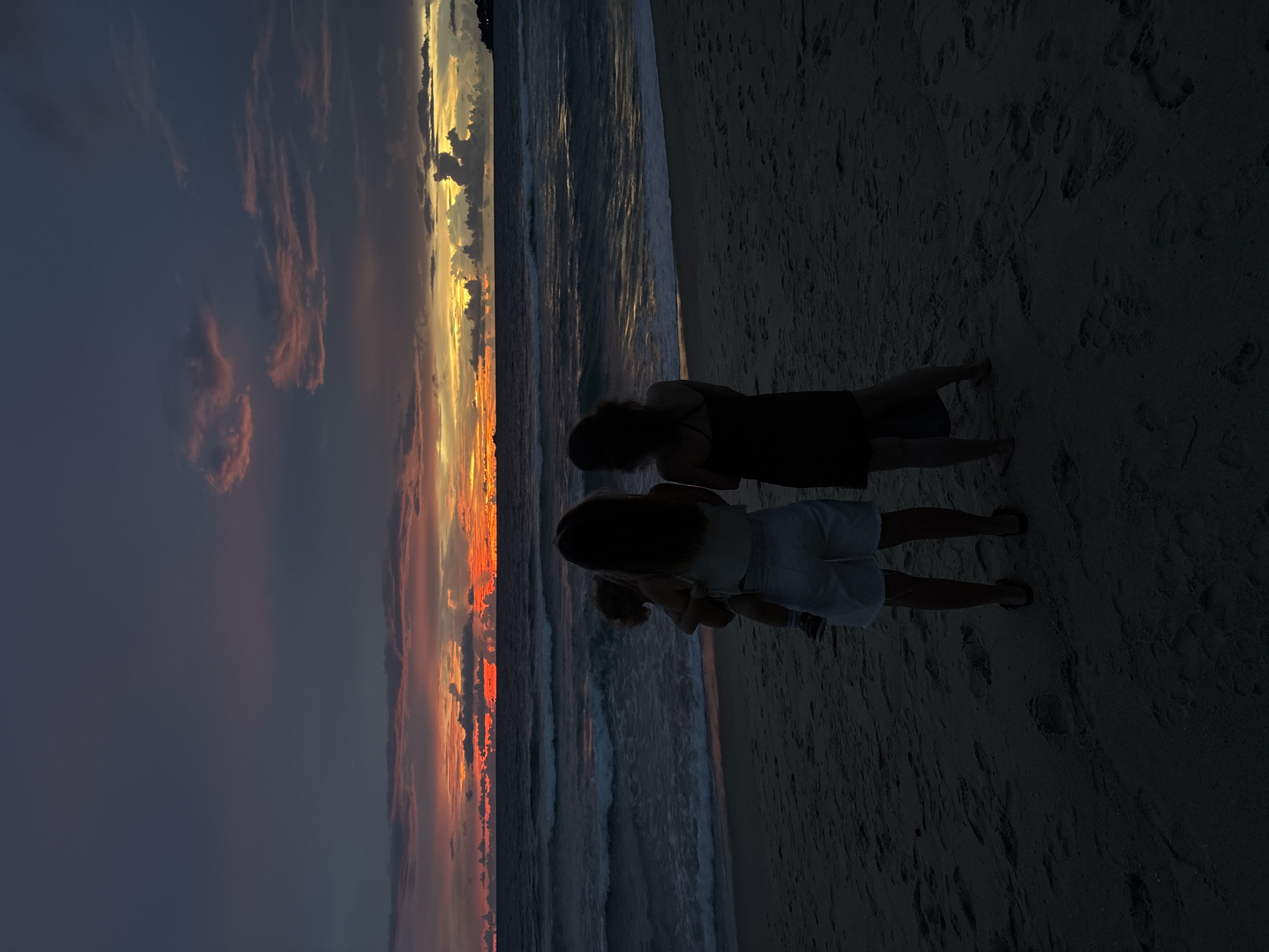 Two females, one holding a child, stand on a sandy beach facing the ocean at sunset. The sky displays vibrant orange and pink hues with dark clouds. Waves gently lap the shore. The image appears to be taken in Choeng Thale, Thailand.