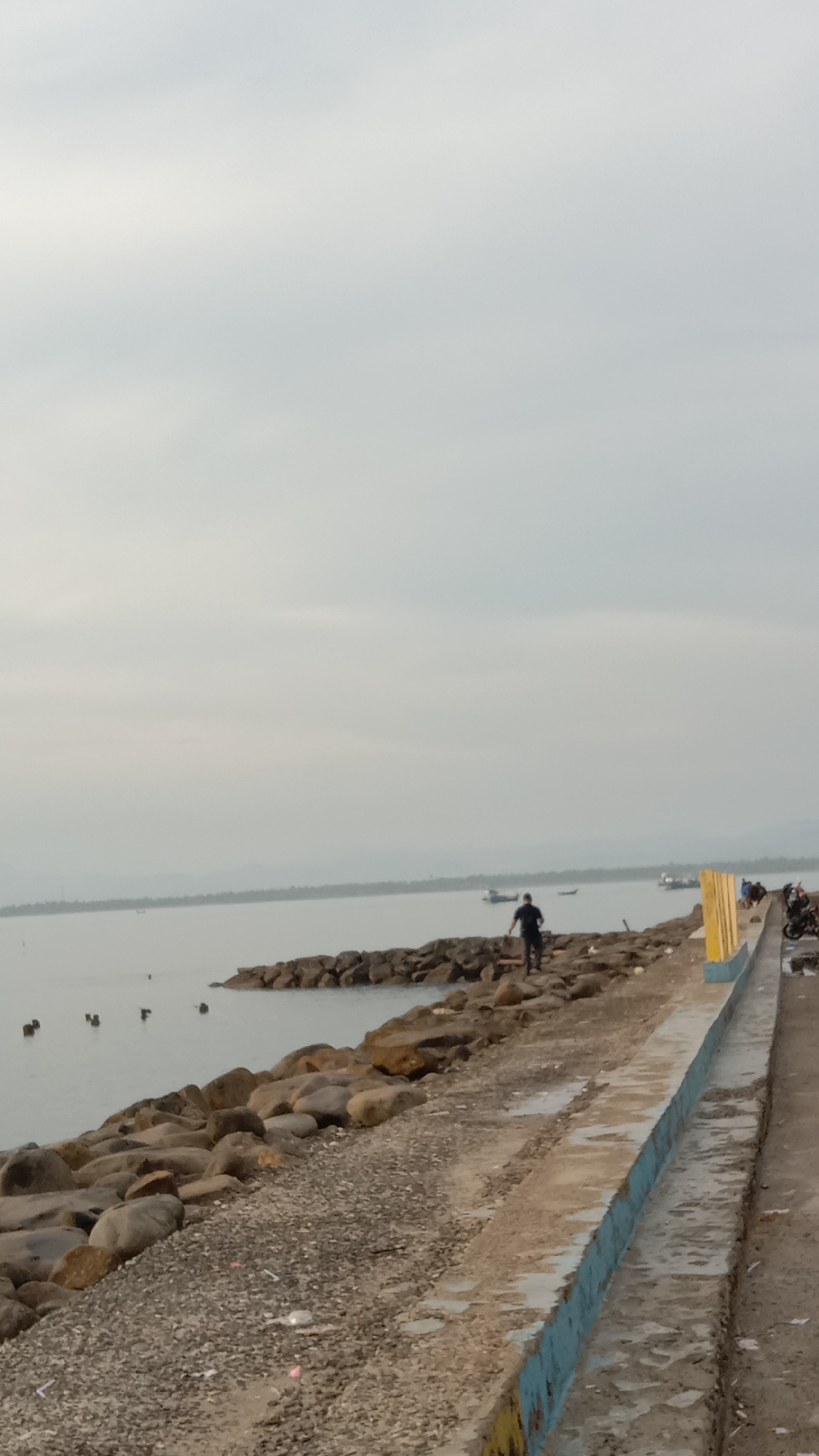 The image shows a coastal scene in Lhokseumawe, Indonesia. A person is walking along a breakwater constructed of large rocks, separating the sea from a concrete walkway. Several boats are visible in the distance. The sky is overcast. Motorcycles are parked near the edge of the walkway. The concrete walkway has a blue painted curb.