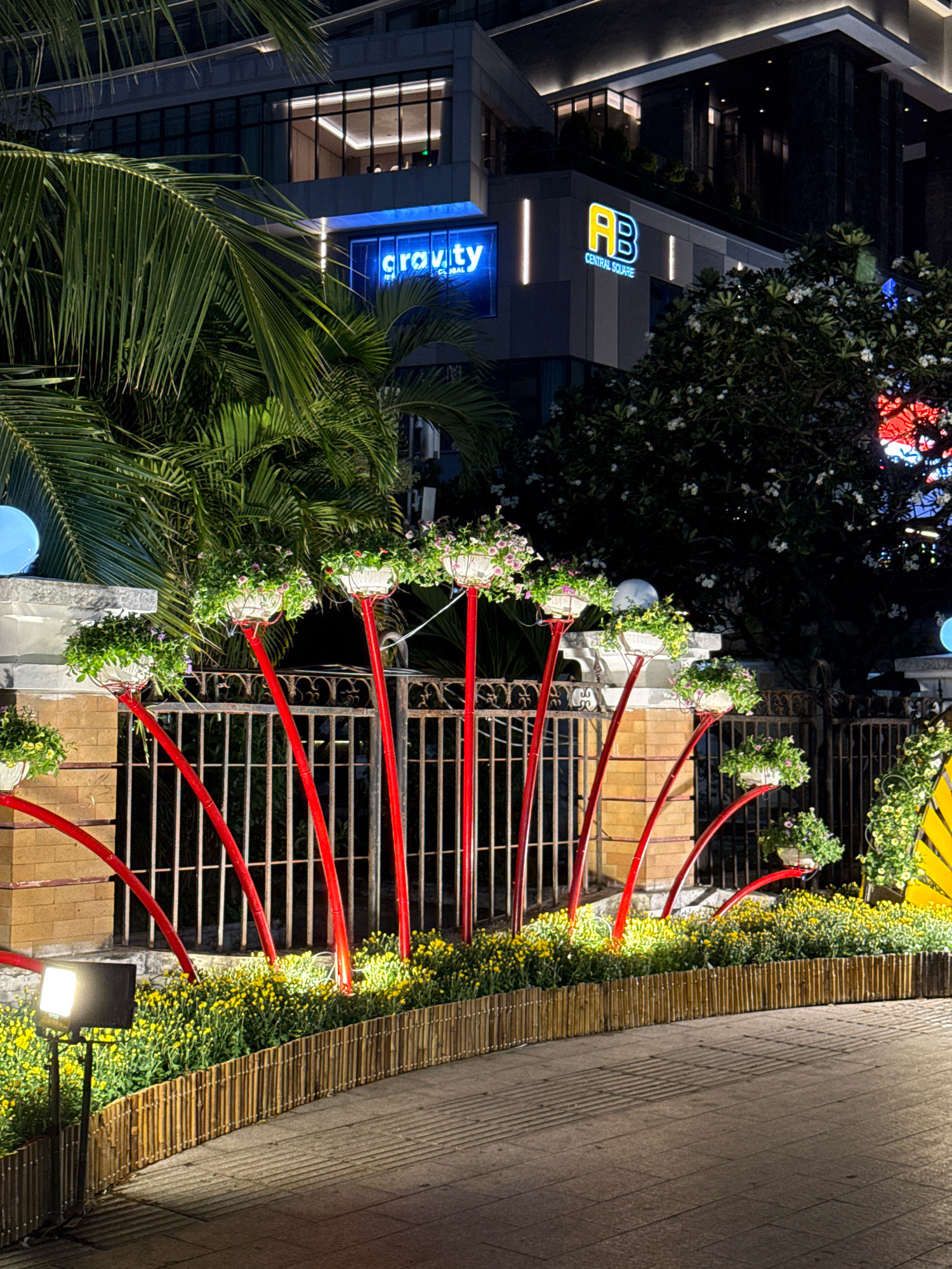 The image depicts an outdoor scene at night, likely in a commercial or urban area. In the background, a modern building is visible, featuring multiple levels of windows and illuminated signage. One sign prominently displays the word "cravity" in blue neon letters, and another adjacent sign shows "AB CENTRAL SQUARE" with yellow and blue neon lettering.
In the foreground, a decorative arrangement of potted plants is displayed. White planters filled with greenery and small flowers are elevated on thin, bright red poles that arch and extend at varying heights. These red poles are interspersed with a black metal fence. Below the fence, a dense bed of small yellow flowers runs along a low bamboo border. A bright floodlight illuminates a section of this floral display and the paved ground in front of it, casting distinct shadows. Palm fronds are visible in the upper left portion of the image, suggesting a tropical or subtropical location. The overall impression is one of nighttime street decoration or landscaping. The signage in the background, specifically "AB CENTRAL SQUARE," strongly suggests the location is in Nha Trang, Vietnam, as this is a known commercial center there.