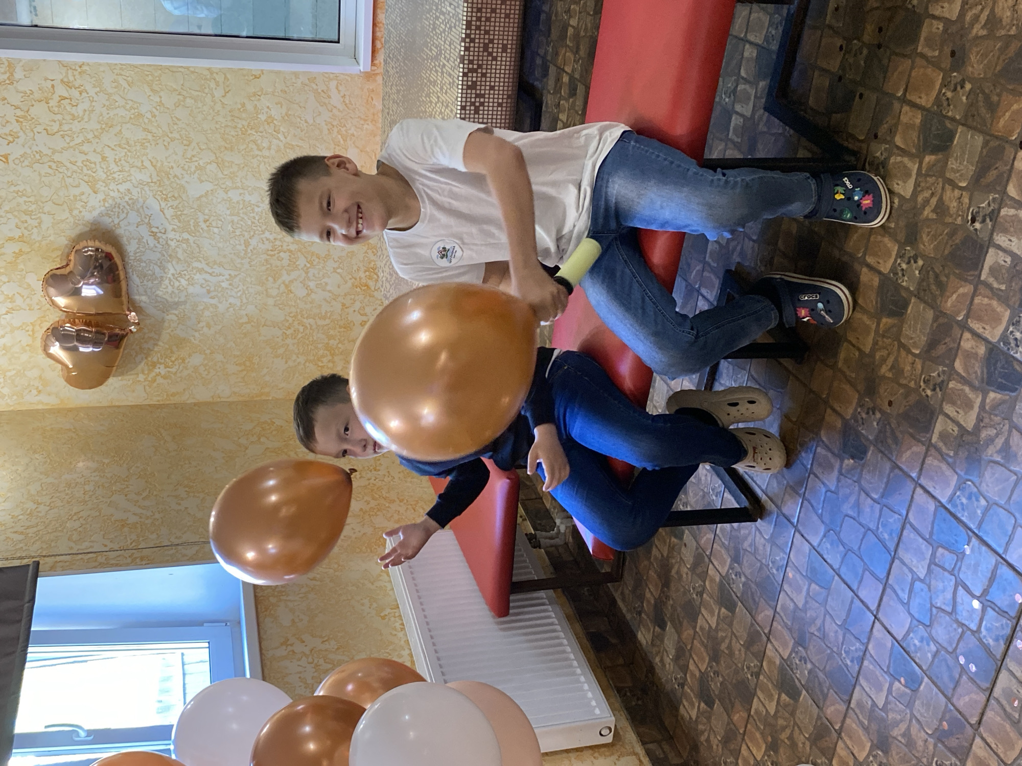 Two boys are indoors, likely at a birthday celebration. One boy, wearing a white T-shirt and jeans, is inflating an orange balloon with a hand pump. He is sitting on a red bench. The other boy, wearing a dark long-sleeved shirt and jeans, is sitting next to him on the bench, watching another orange balloon float. There are multiple balloons, both orange and white, scattered near a radiator. Heart-shaped balloons are attached to a wall. The location is likely an indoor party room. The floor is tiled, and the wall has a textured pattern. The location is in the city of Chaikovsky, Russia.