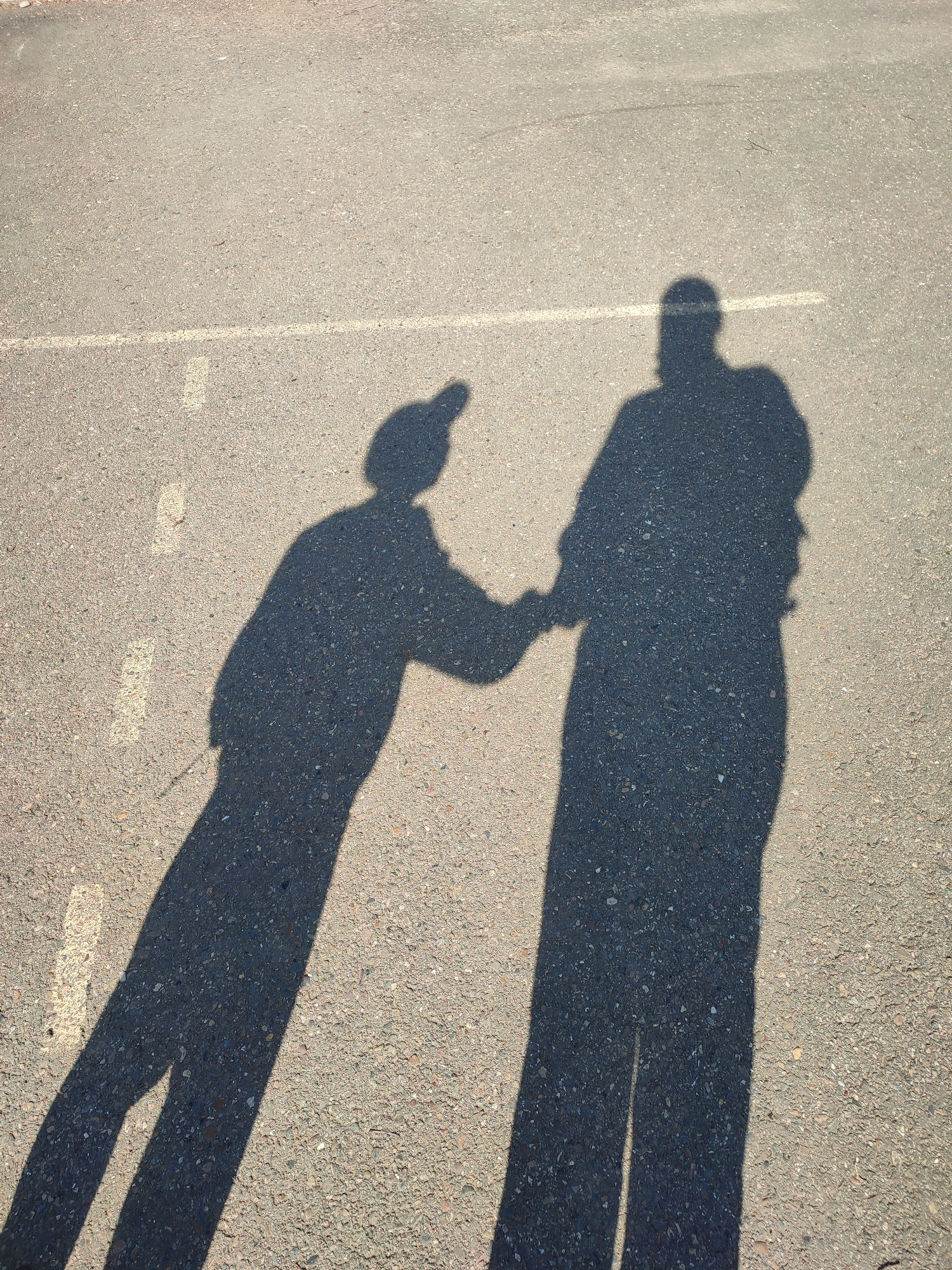 The image shows the shadows of two people on an asphalt surface. One shadow is wearing a hat and has a smaller build, extending their hand towards the other shadow, which is taller and broader. The shadows are cast at an angle, suggesting the sun is at a low angle. The asphalt surface is marked with a few broken white lines. The location, Chaikovsky, Russia, is extraneous to the visual information available in the image.