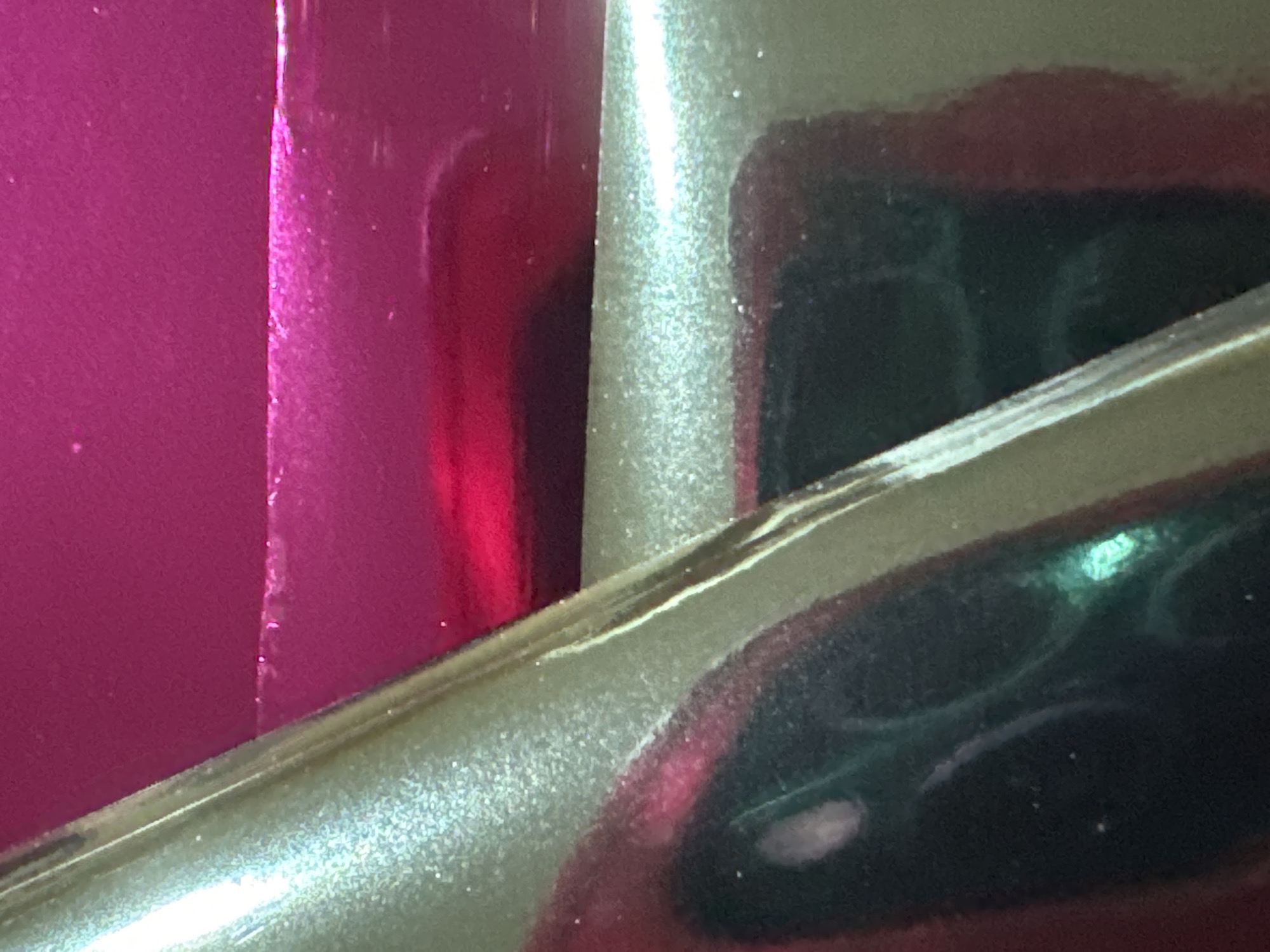 The image depicts a close-up abstract view of a metallic surface with a textured, glitter-like finish. The surface is predominantly a light champagne or beige color and reflects light, creating a shimmering effect. A prominent diagonal line runs across the image, dividing the champagne surface from a deep magenta or fuchsia colored surface. The magenta surface is smoother and appears to be a different material, possibly a highly polished or lacquered finish. There are subtle reflections and highlights on both surfaces, suggesting a glossy and reflective quality. The lighting conditions create areas of bright illumination and shadow, emphasizing the textures and curves of the object. No specific location in the city of Waterford, Ireland, can be identified from this abstract image.