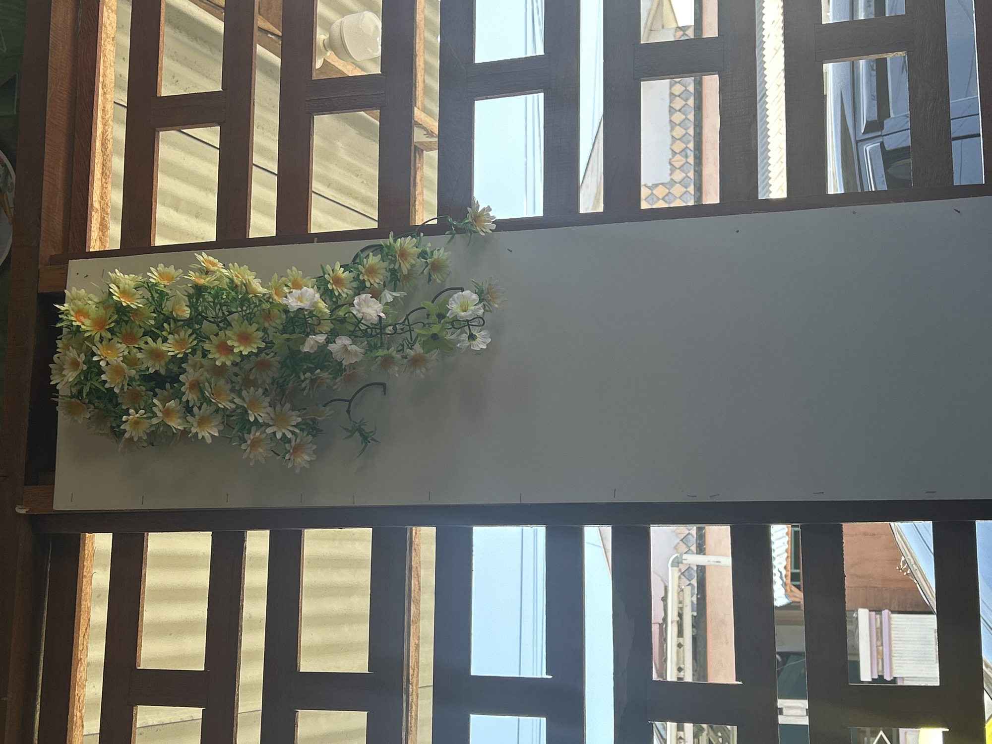 This image captures a section of a building's exterior in Mandaue, Philippines, with a focus on a wall adorned with artificial flowers. The wall is a plain, light-colored rectangular panel, set against a backdrop of dark wooden latticework. This latticework creates a grid of rectangular openings, through which glimpses of the surrounding urban environment are visible.
Through the openings, one can see corrugated roofing materials, suggesting other structures nearby. In the lower portion of the frame, through the wooden slats, parts of a dark-colored vehicle, possibly an SUV, are discernible. The sky appears light blue, indicating daytime. The artificial flowers, a cluster of white and yellow daisy-like blooms with green foliage, are draped downwards from the top of the wall panel, adding a decorative element to the otherwise simple facade. The overall impression is that of an outdoor space, likely a courtyard or alleyway, viewed from a slightly upward angle.