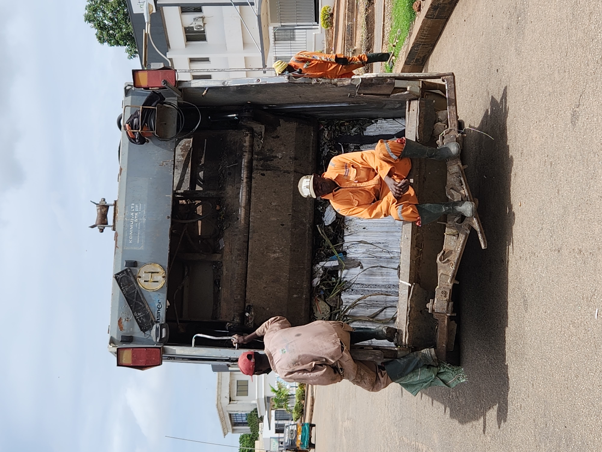 The image captures a daytime scene on a paved street in the city of Unknown, Unknown, featuring a grey rear-loader garbage truck and three sanitation workers. Bright sunlight casts distinct shadows to the left of the truck and workers.
The central object is a heavily used, dirty grey garbage truck, with its rear facing the viewer. The compaction mechanism at the back is open, revealing a load of refuse consisting of foliage, plastic, and other debris. The truck's exterior shows signs of wear, including chipped paint and rust. A red taillight is visible on the upper left rear, beside an empty rectangular license plate bracket and a yellow circular disc marked with a black 'H'. Partial text "KOMMUJA LTI" is visible on the upper panel, and "hsner" is visible lower down.
Three male sanitation workers are depicted.
1. One worker stands on a lower platform at the rear left of the truck, gripping a vertical metal handle. He is seen from behind, wearing a tan long-sleeved uniform, matching pants, a red baseball cap, and dark rubber boots. A green fabric item is partially visible near his left leg.
2. A second worker is seated centrally on the bottom edge of the truck's rear, facing the camera. He wears an orange long-sleeved jumpsuit with reflective stripes, a light-colored baseball cap, and dark rubber boots. His hands are clasped between his knees, and he maintains a neutral expression.
3. A third worker stands on the asphalt road to the right of the truck, facing towards it. He wears an orange long-sleeved jumpsuit with reflective stripes, a yellow knitted cap, and dark rubber boots. He holds a long, thin pole or tool vertically in his right hand.
The street is paved with asphalt and shows minor irregularities. In the background to the left, a light green auto-rickshaw (tuk-tuk) is visible on the street, and white, multi-story buildings with dark-framed windows are present, one bearing a partial "ATM" sign. To the right, additional white, modern-style buildings line the street, one featuring a dark fascia with the partial word "unity." A low curb separates the street from a grassy verge with sparse vegetation. The sky is light blue with scattered white clouds.