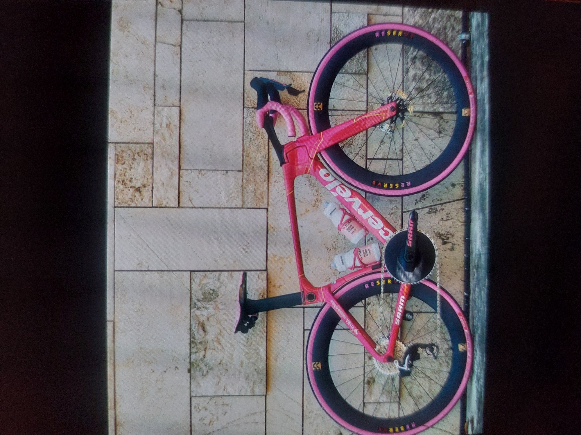 The image shows a pink Cervelo road bicycle leaning against a wall. The bicycle has black wheels with pink tires and SRAM components. Two water bottles are in bottle cages on the frame. The wall behind the bicycle is made of beige-colored stone tiles of varying sizes. A dark, indistinct area is visible on the left and right edges of the image. There is no indication of the location being in Tsarevo, Bulgaria or any specific city. There are no people visible in the image.