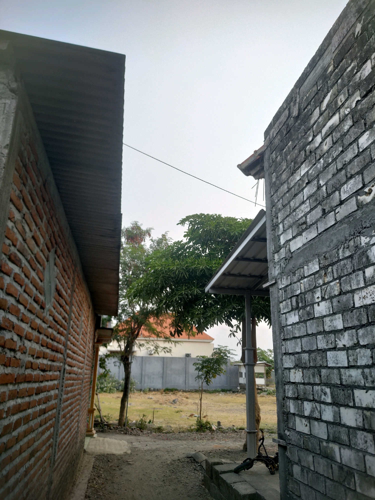 An outdoor, eye-level, medium shot depicts a narrow pathway bordered by two brick buildings, with vegetation and a fence in the background under an overcast sky. The building on the left is constructed with reddish-brown bricks and features a dark, corrugated metal roof. The building on the right is made of weathered, gray and white bricks with visible mortar and a concrete capping. A small, sloped metal awning extends from the gray brick building. Several trees with green foliage are present, including a larger mango tree. In the distance, a light-colored fence runs along a property line, with hints of buildings behind it. The ground is a mixture of dirt and gravel. A partially visible bicycle or similar object rests on concrete blocks near the right building. The scene appears to be in a developing or rural area.