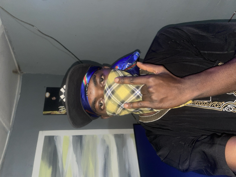 A man with dark skin wears a black hat over a blue and multi-colored head wrap and a yellow plaid mask. He is posing indoors and giving the "rock on" hand sign. He wears a black shirt with white patterned embroidery along the neckline. In the background is an abstract painting with grey and white colors. Another, smaller, framed picture hangs on the wall behind him, near a light fixture. The room's walls are a muted grey-blue.