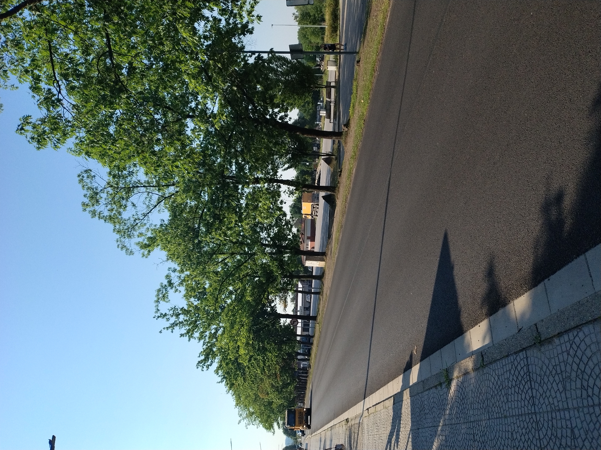 The image shows a street scene in Hluboká nad Vltavou, Czechia, on a clear day. A yellow truck is driving on the right side of a paved road lined with trees. A sidewalk with a decorative pattern runs along the left side of the road. In the background, there are buildings and more trees. A person is visible walking on the right side of the road in the distance. The scene appears to be a typical street in a town or city.