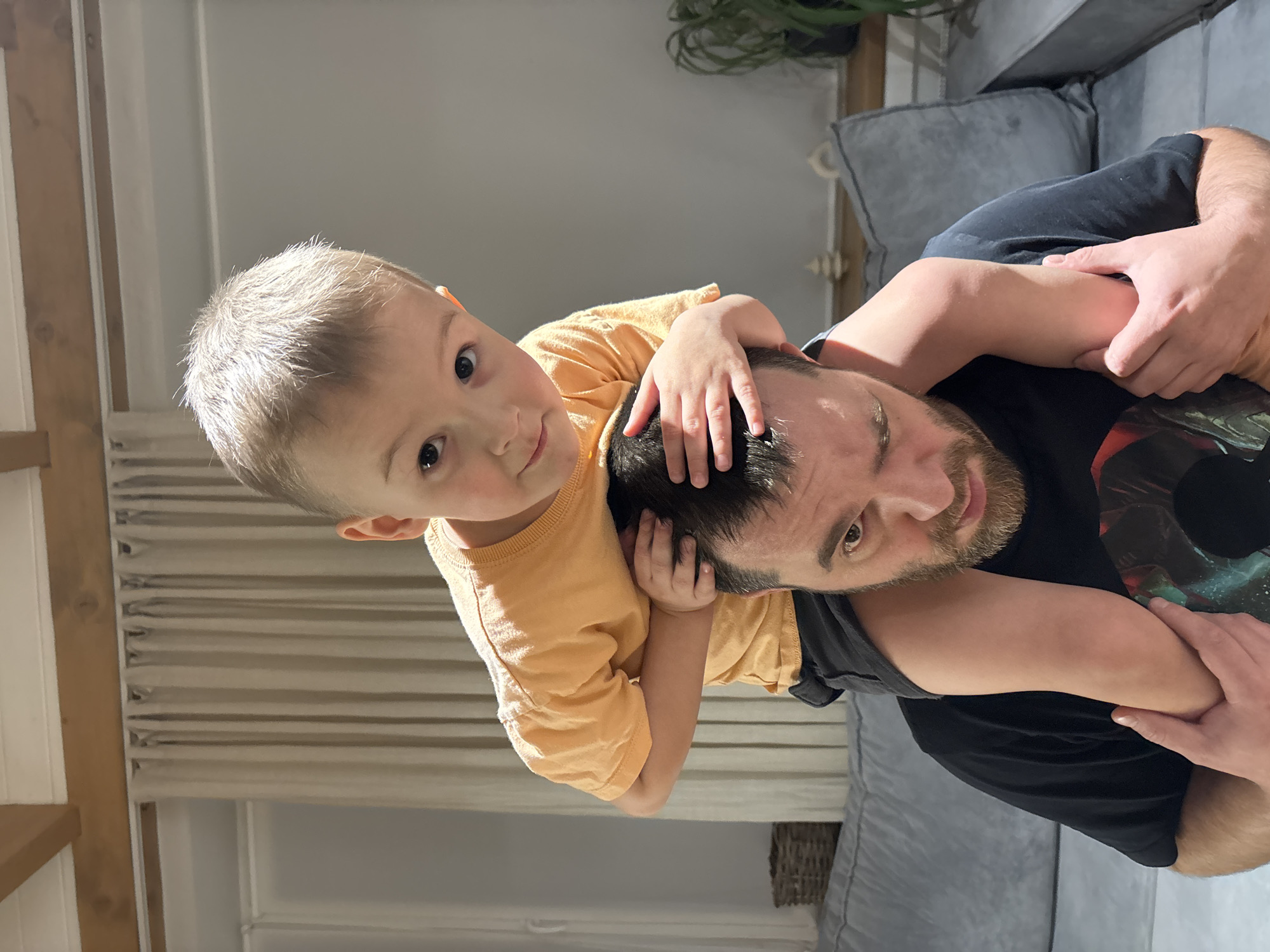 The image, oriented correctly, depicts a heartwarming indoor scene featuring a man and a young child in Chaikovsky, Russia. A fair-skinned man with short dark hair and a beard, wearing a dark t-shirt, is positioned in the lower right of the frame, looking towards the camera with a gentle expression. A young boy, likely blonde, with bright eyes and wearing a light orange t-shirt, is playfully perched on the man's shoulders or upper back. The child's face is turned directly towards the camera, and his hands are gently placed on the man's head, suggesting a close and tender interaction.
The setting appears to be a well-lit interior, possibly a living room or common area of a home. In the background, to the right, a grey upholstered sofa or couch is visible, along with a glimpse of a green potted plant with curly leaves above it. A partially visible wicker basket sits on the floor near the sofa. To the left, there's a light-colored wall with horizontal blinds or curtains, and a prominent wooden beam or pillar. The lighting suggests it is daytime, with highlights visible on the child's hair and the man's skin. Both individuals seem calm and content, conveying a sense of familial affection and playfulness. There is no visible text in the image.