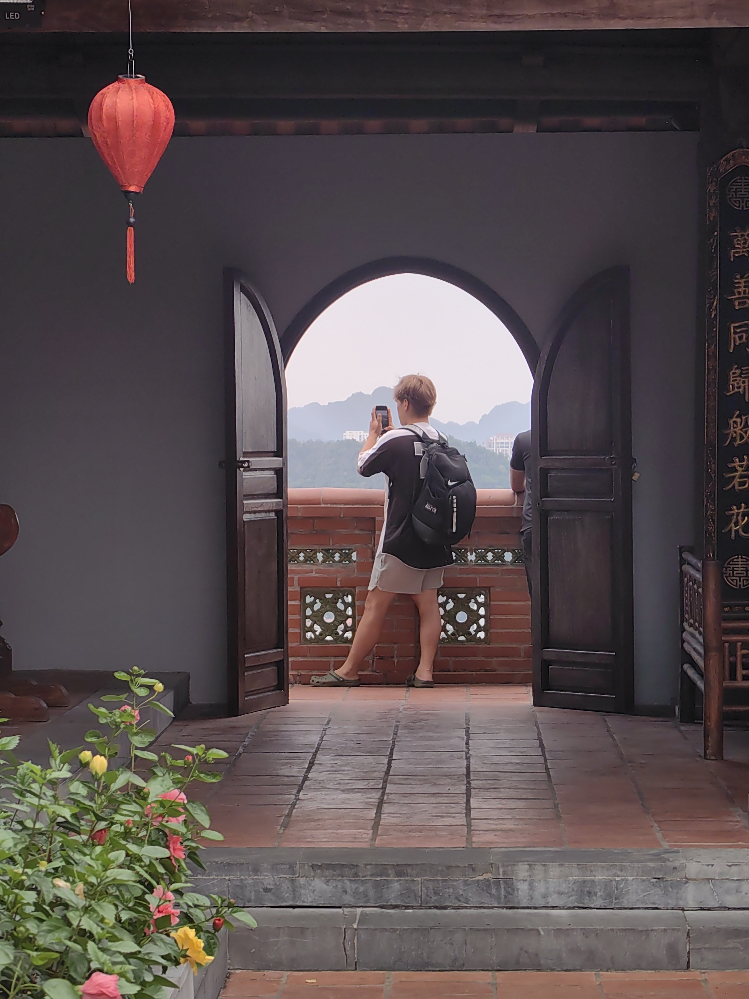 A photograph captures a person standing at an arched opening in a building, seemingly taking a photo of a distant landscape with a phone. The landscape features mountains in the background. The person is wearing casual clothing, including shorts, a black and white shirt, and sandals. A black backpack is on their back. The opening is flanked by open wooden doors. A red lantern hangs from the ceiling. To the left, there's a plant with pink and yellow flowers. To the right, an Asian-style piece of furniture is visible. In the upper left, the letters "LED" are visible. Writing in an East Asian language is on the right side of the image.
The image was taken in Hong Gai Ward, Vietnam.