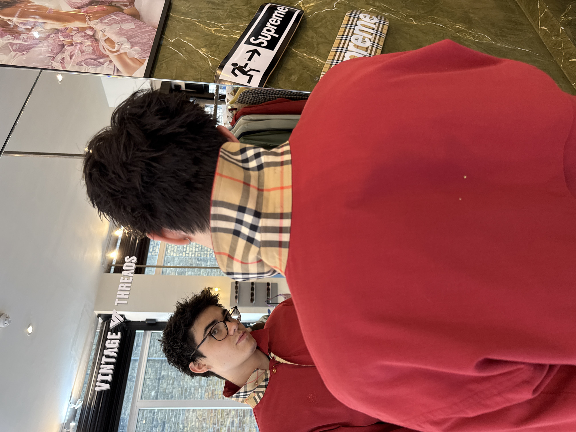 A person wearing a red Burberry sweatshirt with a plaid collar stands inside the Vintage Threads store in London, United Kingdom, looking at their reflection in a mirror. The store's logo is visible in the mirror, and two Supreme skateboards are displayed on a shelf above the person's head. A framed picture is leaning on the shelf. The store has light-colored walls and flooring.