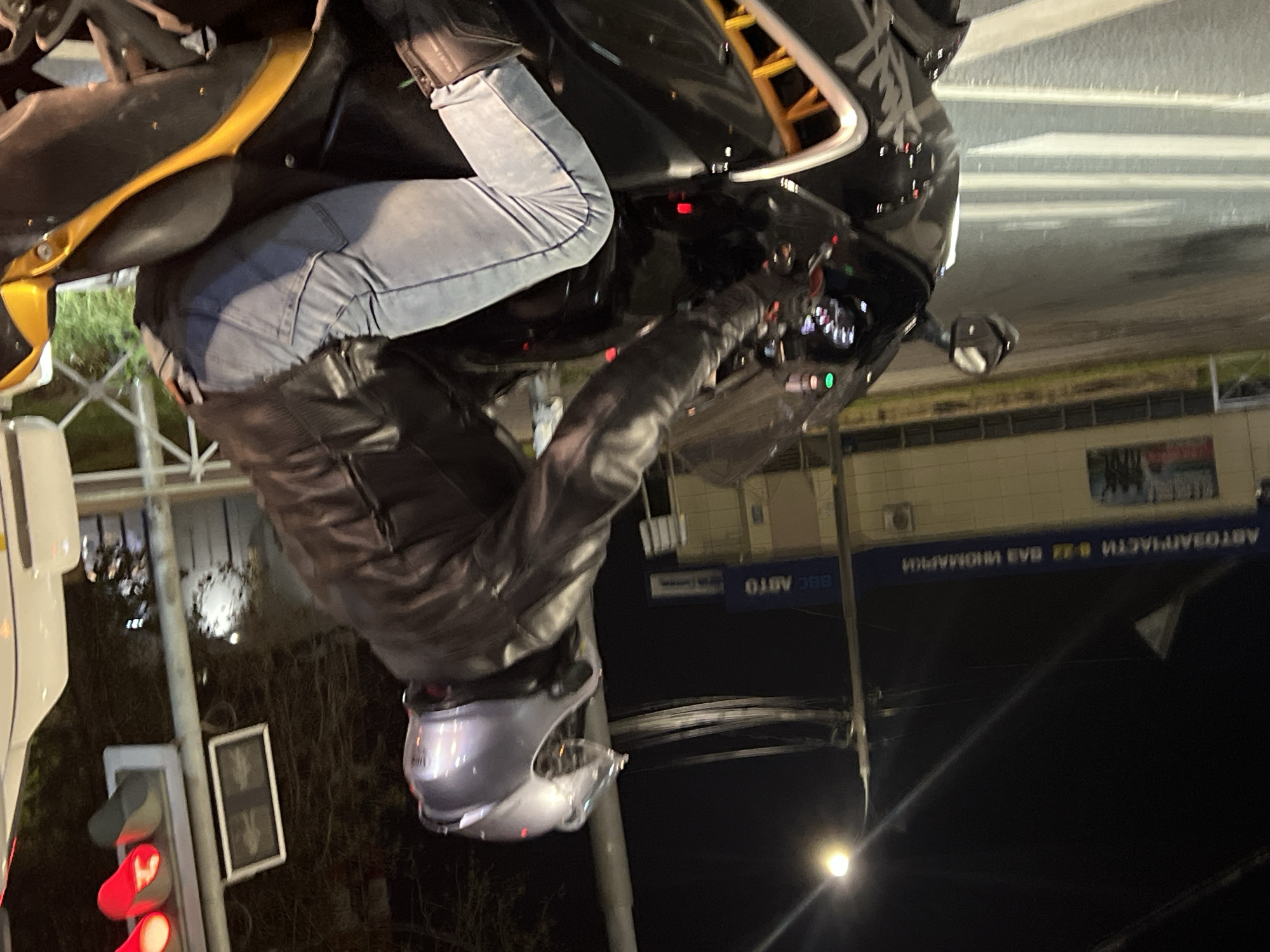A nighttime exterior shot captures a person on a black motorcycle at a crosswalk in Klin, Russia. The individual is wearing a silver helmet, a dark leather jacket, and light blue jeans. The motorcycle's dashboard lights are illuminated, and the taillight is red. A traffic light above and to the right of the motorcycle displays a red signal. In the background, a building with blue signage that reads "АВТОЗАПЧАСТИ 24/7 ВАЗ ИНОМАРКИ" (Auto Parts 24/7 VAZ Foreign Cars) is visible, along with streetlights and overhead wires. The ground is wet, suggesting recent rain. The scene is dimly lit, with artificial light sources casting shadows.