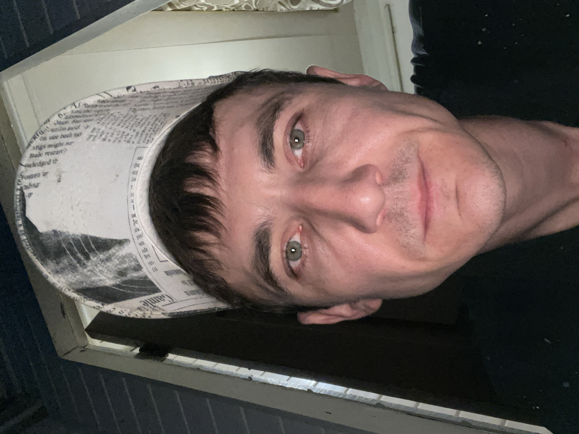 A man wearing a cap with a newspaper print pattern is looking directly at the camera. He has light skin, dark brown hair, and green eyes. He is wearing a black shirt. The background appears to be an indoor setting with a doorway and a curtain. The image was taken in Laishevo, Russia.