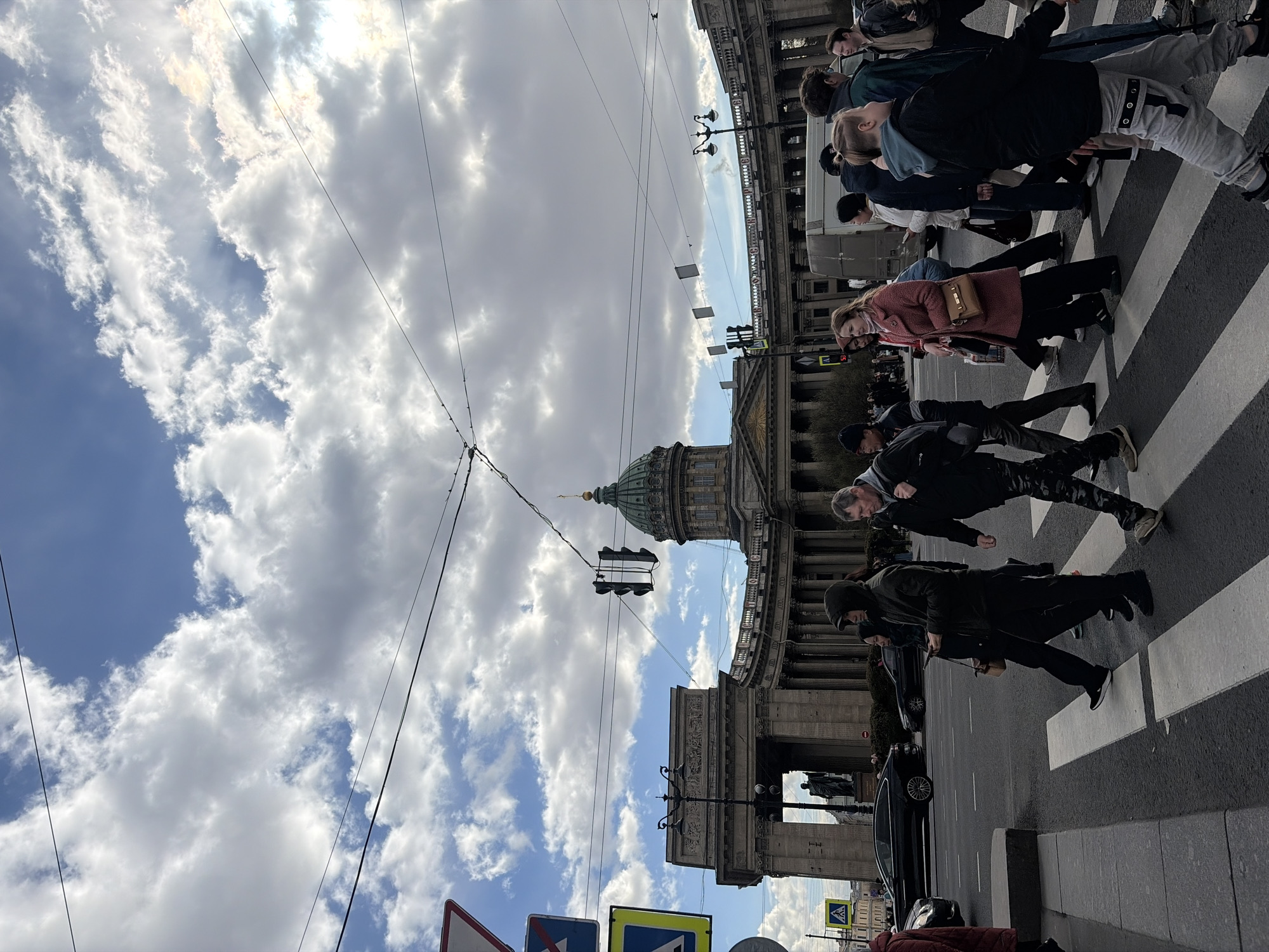 The image depicts a bustling street scene in Saint Petersburg, Russia, with the iconic Kazan Cathedral prominently featured on the right. Numerous pedestrians, dressed in casual clothing, are actively crossing a zebra crossing from the left towards the right side of the frame.
The Kazan Cathedral, a magnificent Neoclassical building, dominates the right half of the image with its large green dome, grand colonnade, and ornate facade. Overhead, power lines and streetlights crisscross the sky. The sky itself is partly cloudy, with bright white and grey clouds interspersed with patches of clear blue, indicating a bright, sunny daytime.
In the foreground and middle ground, people of varying ages are seen walking. A black car is visible on the street, near the pedestrian crossing. Several road signs are present: a blue square sign with a white arrow pointing straight, and a yellow diamond sign also with a black arrow pointing straight. The overall scene captures everyday urban life in front of a significant architectural landmark.