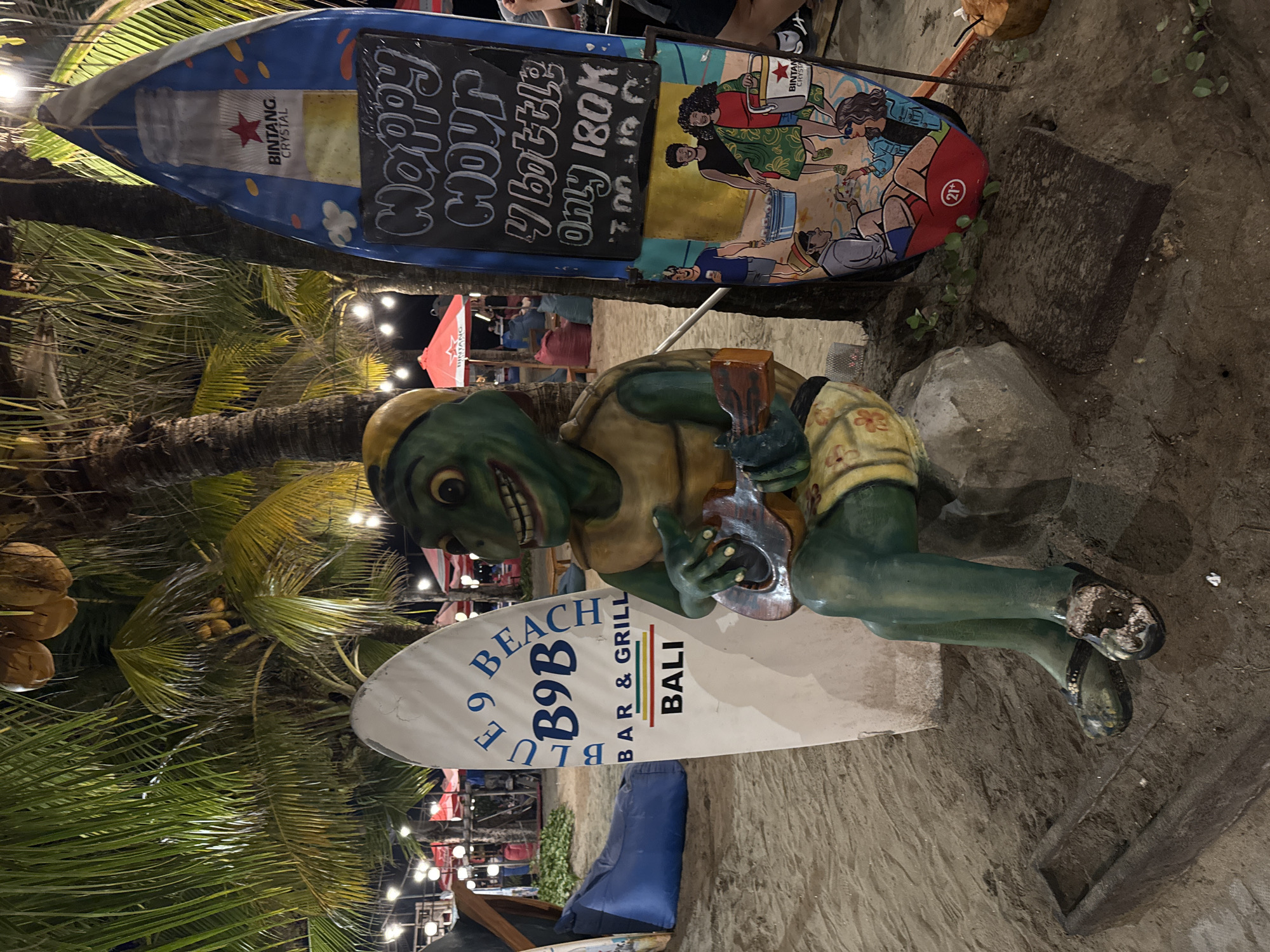 In Kuta, Indonesia, a lively beach scene unfolds at night, illuminated by strings of warm lights. A prominent surf-shaped sign, emblazoned with "BINTANG CRYSTAL," stands to the right of a whimsical, green, anthropomorphic turtle statue. The statue, depicted playing a small ukulele, is adorned in a yellow cap and patterned shorts, seated on a rock. Beside it, a white surfboard-shaped sign advertises "BLUE 9 BEACH BAR & GRILL BALI." Further to the right, a black chalkboard sign announces "HAPPY HOUR 4 bottles Only 180K 7.00 - 10.00 PM." The background reveals more of the beach atmosphere, with red umbrellas and beanbag chairs scattered across the sand, hinting at a bustling establishment. The overall impression is a cheerful and inviting spot for evening relaxation and entertainment on the beach.