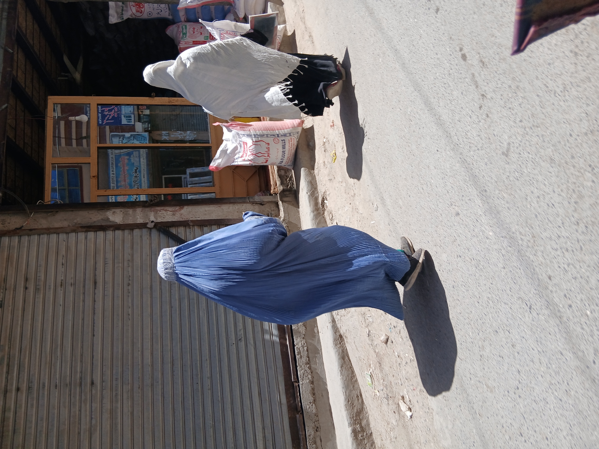 This media file depicts a street scene in Quetta City Tehsil, Pakistan, during daylight hours with strong sunlight casting distinct shadows.
Two individuals, likely women, are walking away from the camera on a paved street. The person in the foreground wears a pleated, bright blue full-body burqa with a white, netted face covering. The person behind her is clad in a white head covering (hijab) and a black garment, featuring a striped black and white pattern on the sleeve.
The street is paved with asphalt or concrete, showing some dirt and debris along the edges. To the left, a building features a large, closed corrugated metal roll-down shutter. To the right, a wooden-framed shop window displays various items and reflections, including a visible light bulb. Above this window, several large sacks are stacked; some are white, and one is reddish-pink.
Visible text on a white sack reads "DUA FLOUR MILLS", "AF", and Urdu script including "جناب", "سارق آباد", and "جنرل ملز". A reddish-pink sack displays "الرحمن" (Al-Rahman). Text reflected or displayed in the shop window includes "NOW", "alrow saal", "۱۷۹", "90 17372 & MUNIMOJA JAN", and mirrored Urdu text such as "آقا" and "یران میشد".