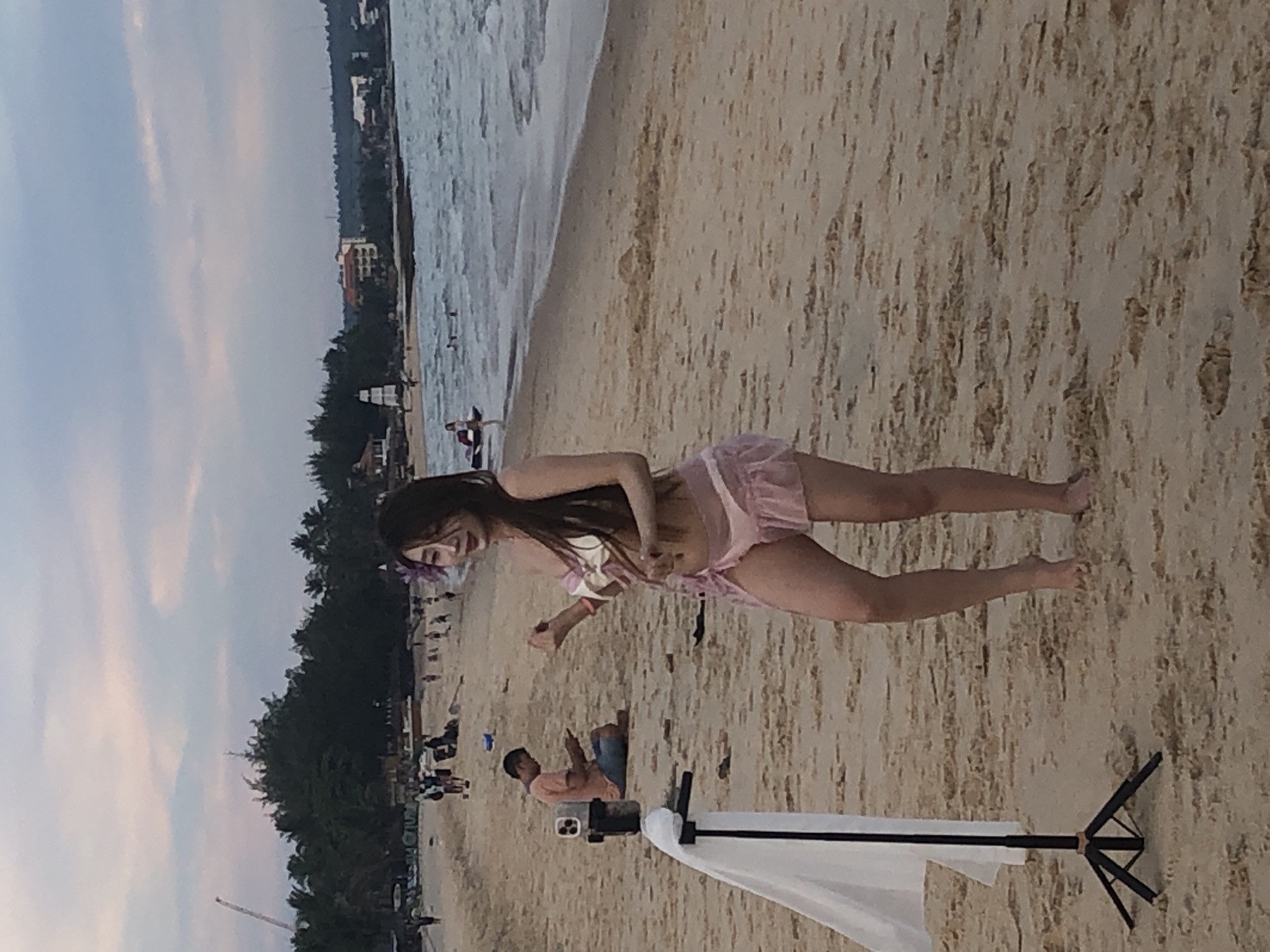 A young woman in a pink bikini poses on a sandy beach with the ocean in the background. She has long, dark hair and a flower in her hair. She is holding her hands up and appears to be dancing. A tripod with a white cloth draped over it stands to her left. In the background, a man sits on the sand looking at his phone. Further back, other beachgoers are visible, along with palm trees and buildings along the coastline. The sky is overcast with soft, diffused light. The location is identified as Phu Thuy Ward, Vietnam.