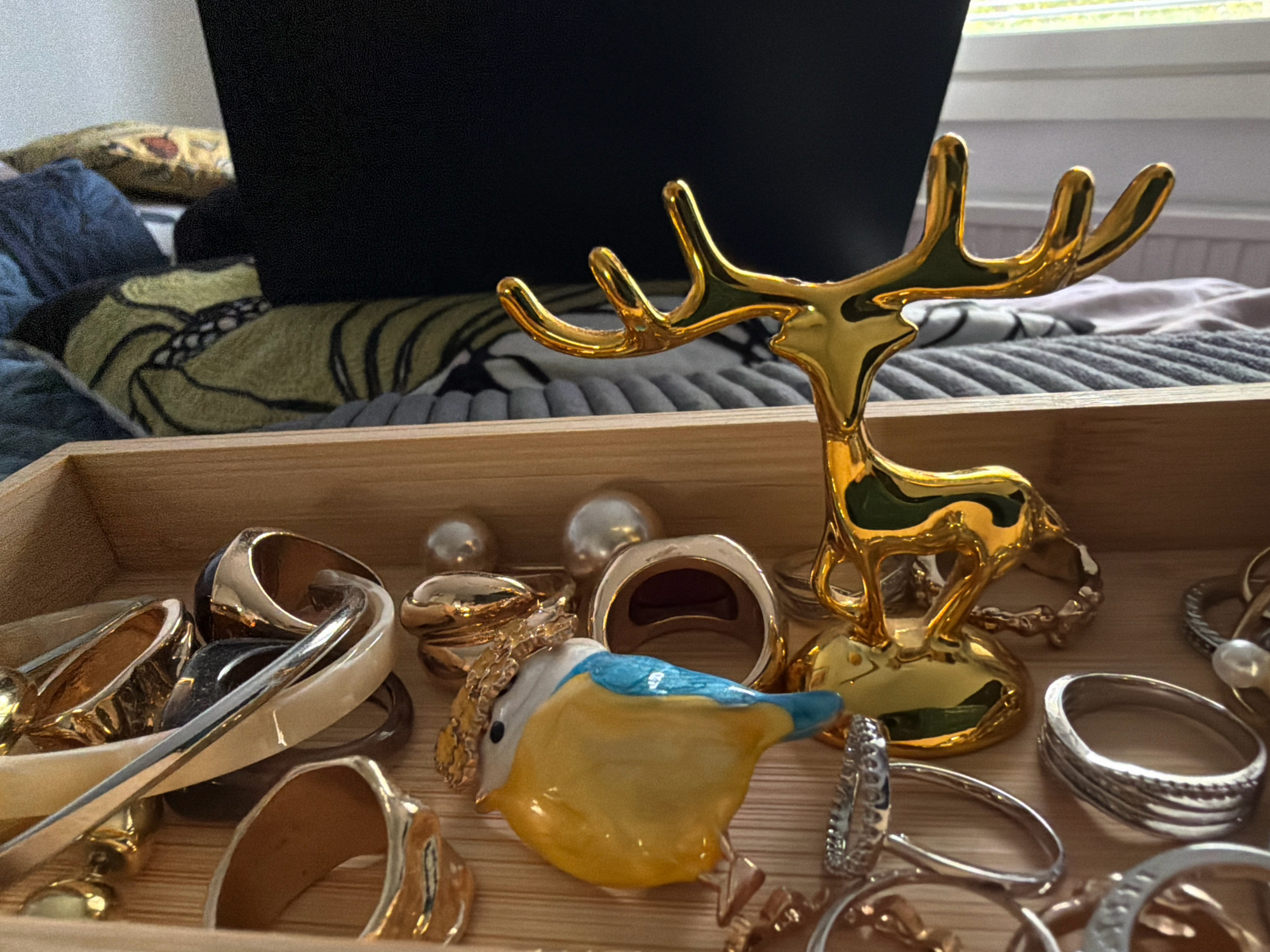 A close-up, eye-level shot shows a wooden tray filled with jewelry and a decorative golden deer figurine. The jewelry includes several rings, some with intricate designs and some with pearls. A small, colorful ceramic bird figurine is positioned in the center of the tray. The background is blurred, but appears to be a bedroom or living area with pillows and blankets. The lighting suggests an indoor setting, possibly during the daytime. The scene is static, with no apparent actions or interactions occurring. The image does not provide any information about a specific location in Helsinki, Finland.