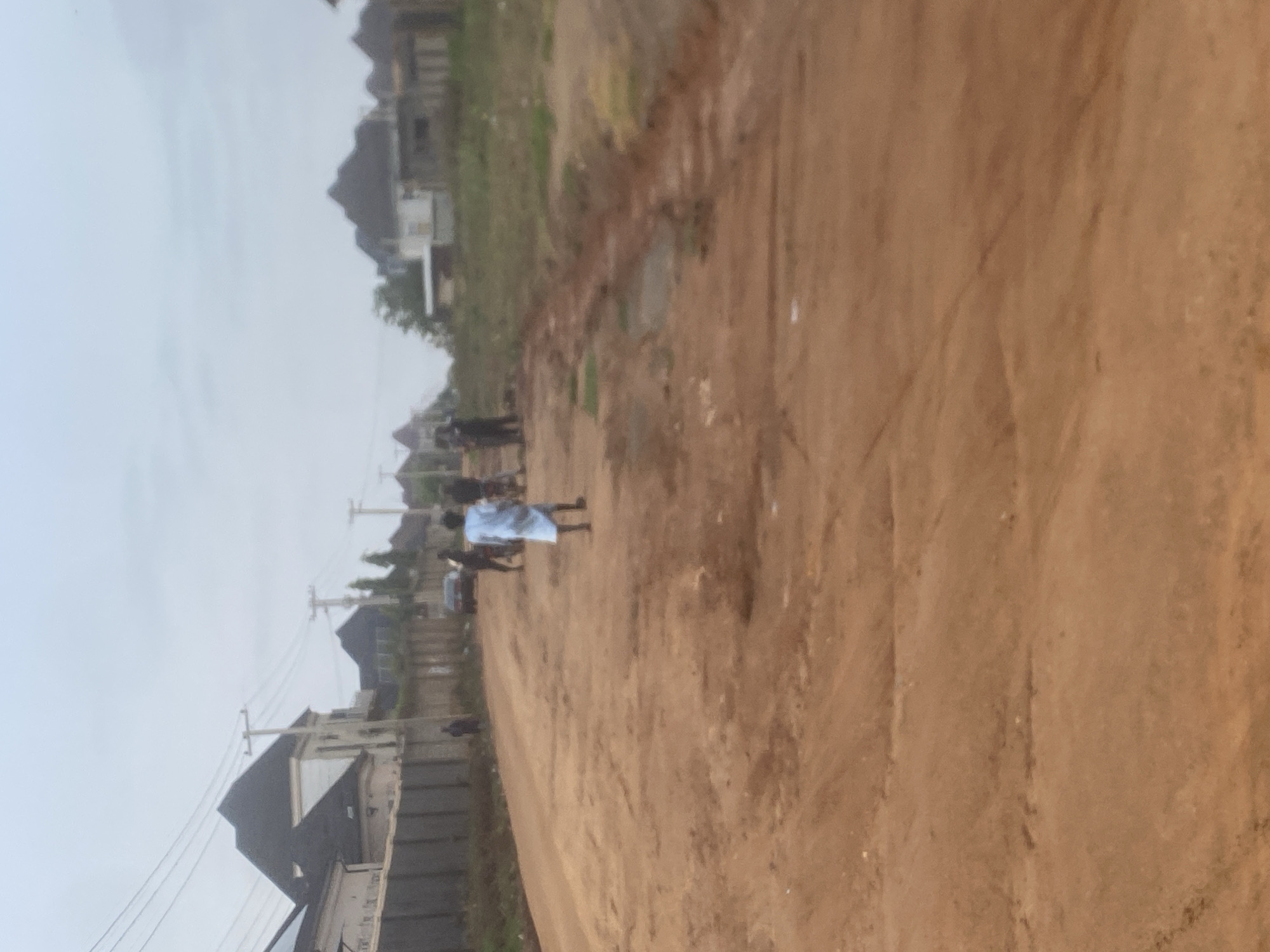 The image depicts a dirt road in Iddo Tudunwada, Nigeria. Several individuals are present on the road. One person, wearing a light blue garment, stands in the center foreground. To the left and further down the road, a dark-colored car is visible. The road is unpaved and appears to be under construction or in a state of disrepair, with uneven surfaces and patches of mud. On either side of the road, there are houses with dark roofs and fences. Electrical poles and wires are also present in the background. The sky is overcast, suggesting a cloudy or hazy day.
