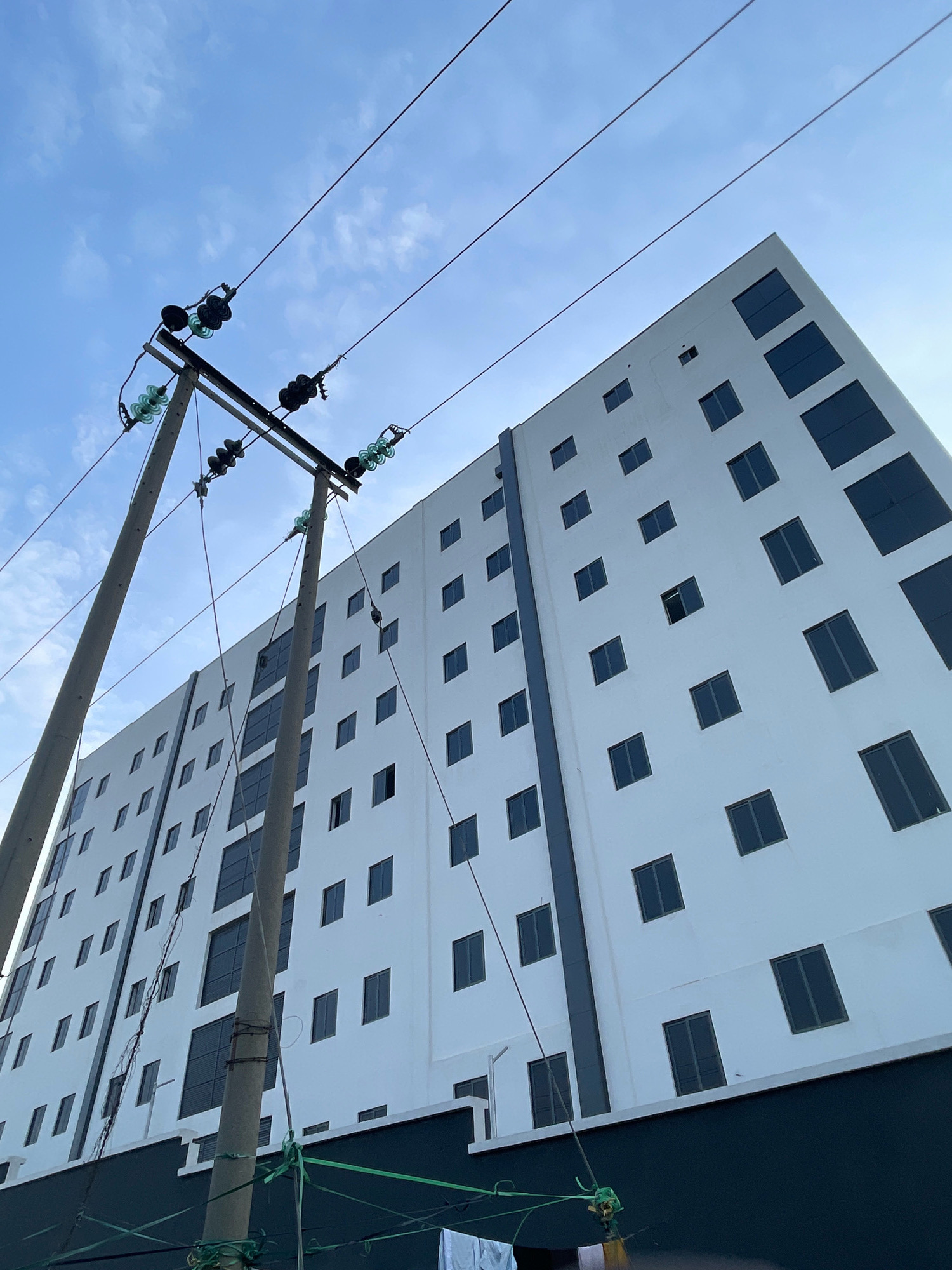 An exterior, low-angle, full shot captures a modern, white, multi-story building in Abuja, Nigeria. The building features numerous rectangular, dark-tinted windows arranged in a grid pattern across its facade. In the foreground, two utilitarian concrete utility poles with electrical wires and insulators stretch diagonally across the frame, contrasting with the building's sleek architecture. The sky above is a light blue with wispy white clouds. The bottom edge of the image shows a dark, textured lower section of the building and some hanging white laundry.