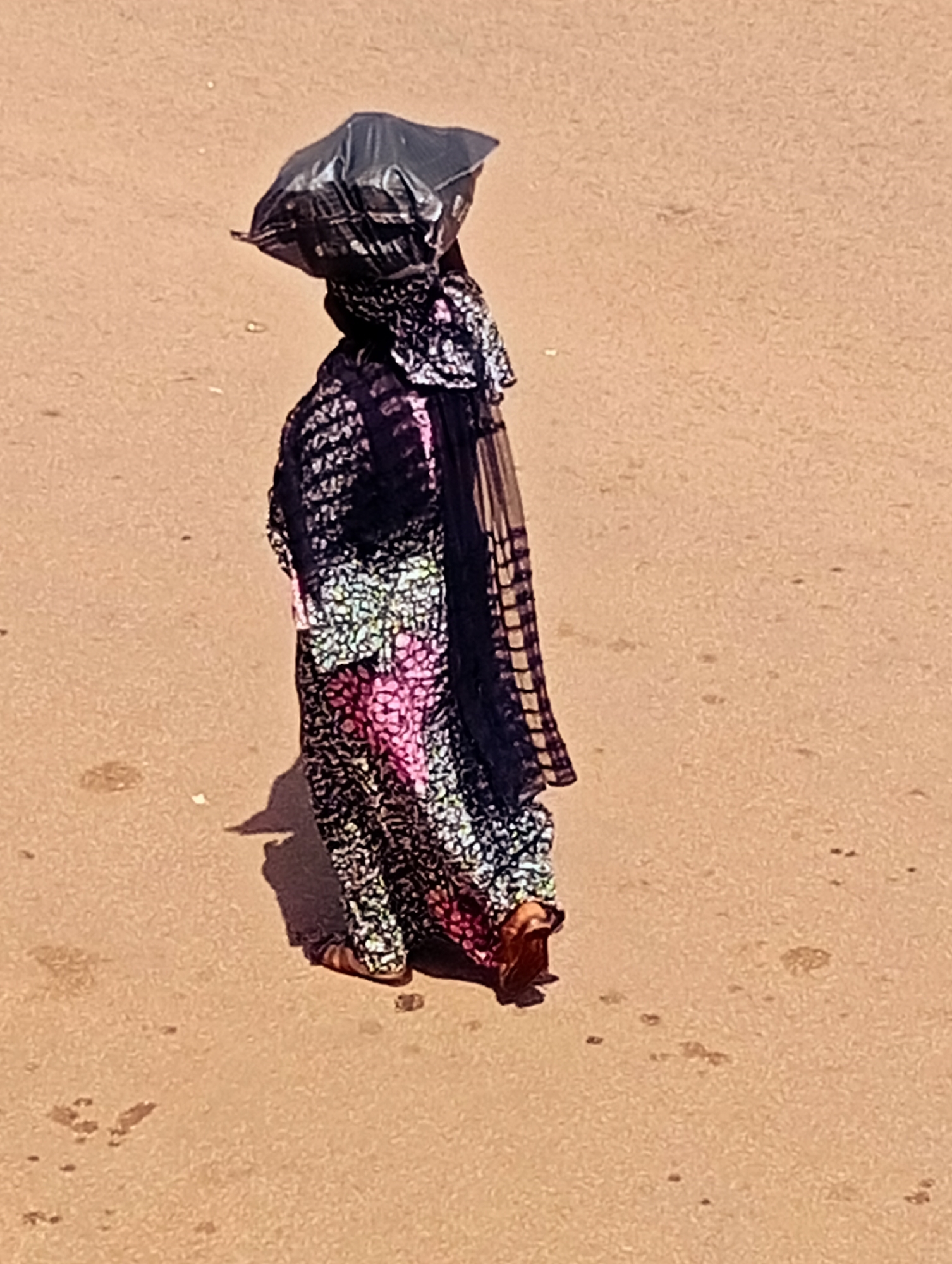 A person walks along a dusty road in Bukuru, Nigeria, carrying a black plastic bag balanced on their head. The individual is wearing a long, colorful patterned dress and a headscarf. Their feet are visible, wearing sandals. The ground is a light brown, sandy color, with some small dark marks and debris scattered across it. A shadow is cast by the person on the ground.