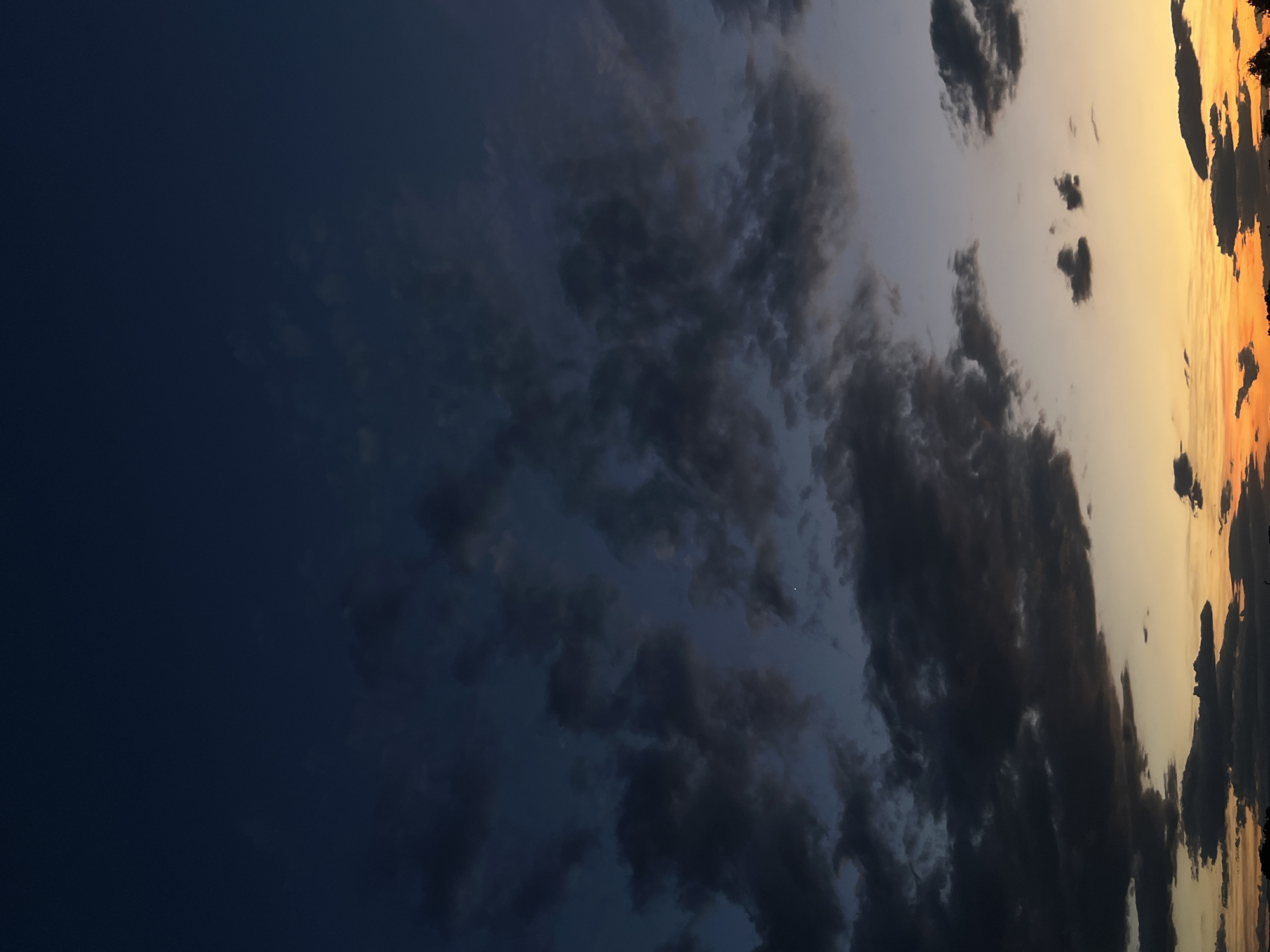 The image shows a sunset over the city of Plovdiv, Bulgaria. The sky is a deep blue, with streaks of orange and yellow from the setting sun. Dark clouds are scattered across the sky, and a faint star or planet is visible. The horizon is lined with trees and buildings.