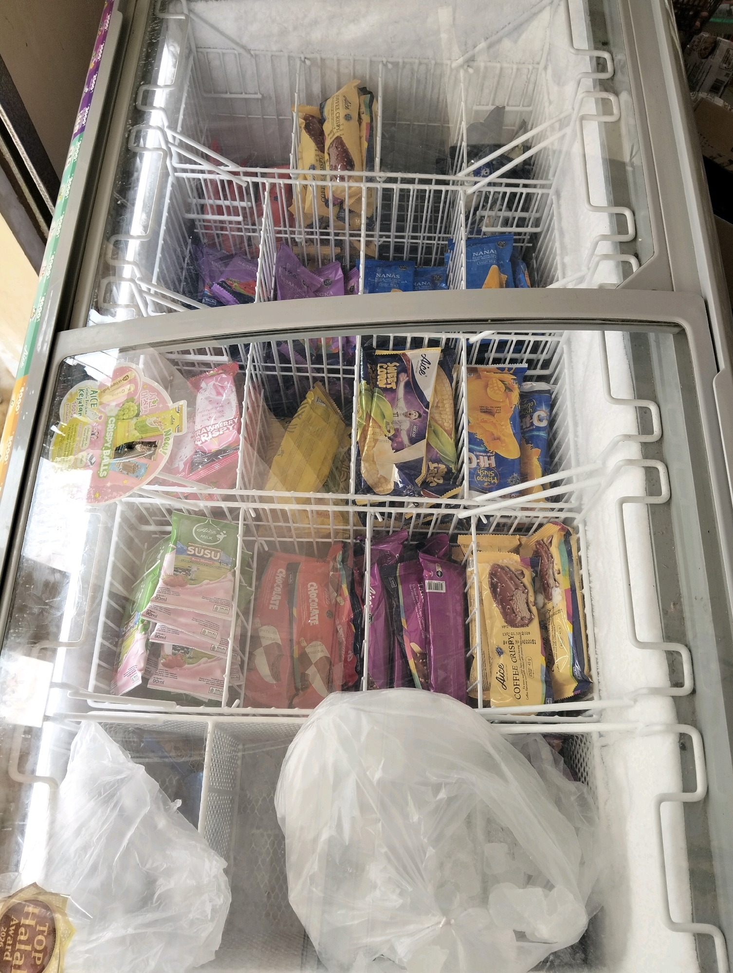 An interior view of a commercial freezer displays an assortment of frozen food items, predominantly ice cream bars and packaged desserts. The freezer is divided into multiple shelves and compartments, each stocked with various brands and flavors. In the top shelves, yellow and purple packaged ice cream bars are visible, some featuring images of fruit and chocolate. The middle shelves contain more ice cream products, including those with labels indicating "Strawberry Crispy," "Mango Slush," and "Chocolate." Several packets of what appear to be milk drinks or yogurts are also present, labeled "SUSU" and "HALAL." The bottom section of the freezer holds plastic bags, one of which is transparent and filled with ice cubes, indicating the freezer is in operation. The environment is cold, with visible frost accumulating on the interior surfaces of the freezer. There are no people or discernible actions depicted in the image. The overall scene suggests a retail or storage setting for frozen goods. The specific location within Patrol, Indonesia, cannot be determined from the visual information provided.