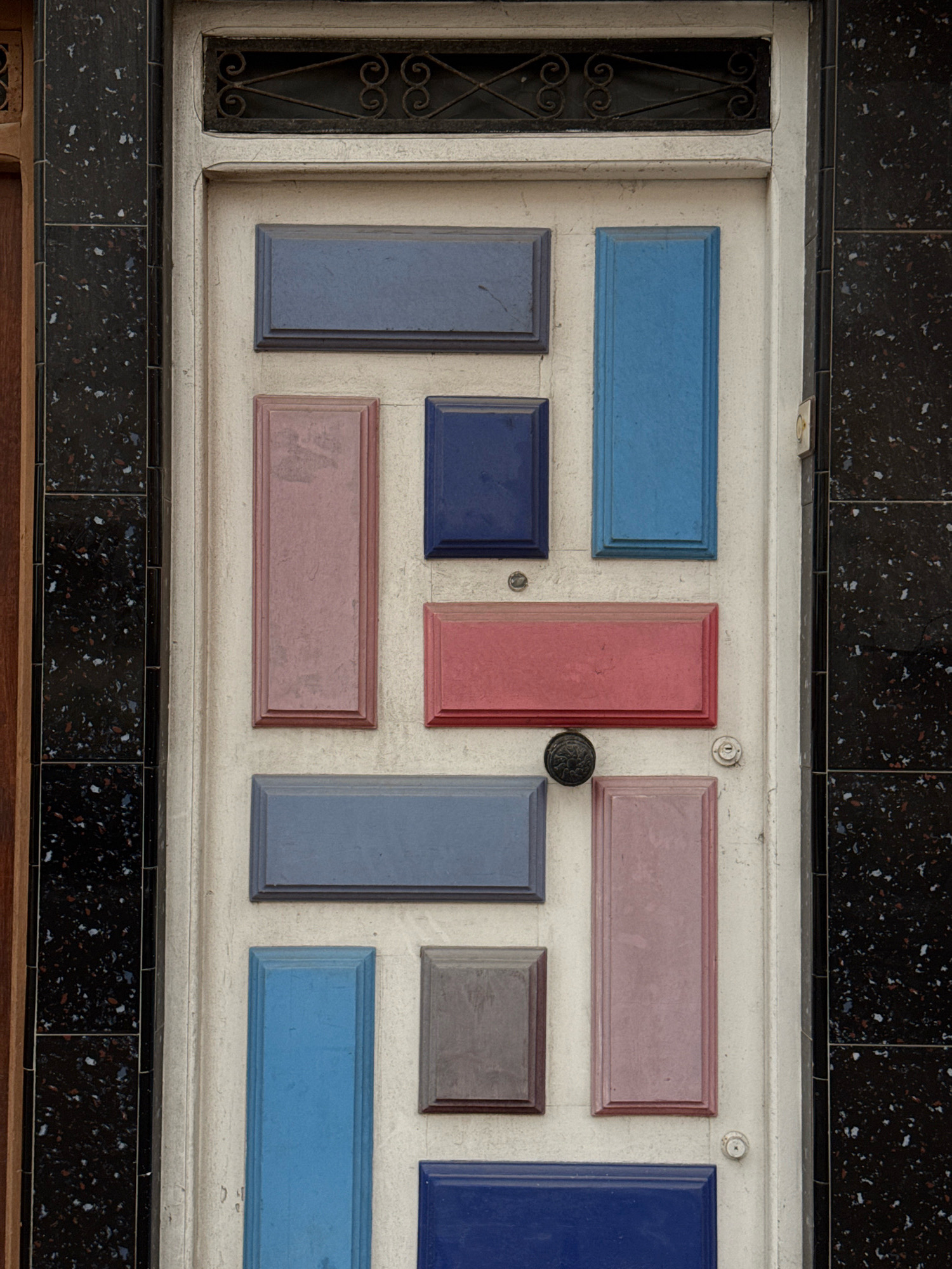 The image displays a close-up of a white door in Essaouira, Morocco. The door is adorned with an array of rectangular and square panels in varying shades of blue, pink, and red, arranged in a geometric pattern. These panels are set into the door's surface, creating a textured and visually striking design. A dark, ornate doorknob is centrally located, and a small keyhole is visible below it. The door is framed by a dark, speckled architectural element on the right and a wooden element on the left. Above the door, a decorative metal grate with a scrolling pattern is visible. The overall impression is one of unique, artistic decoration applied to a functional entryway.