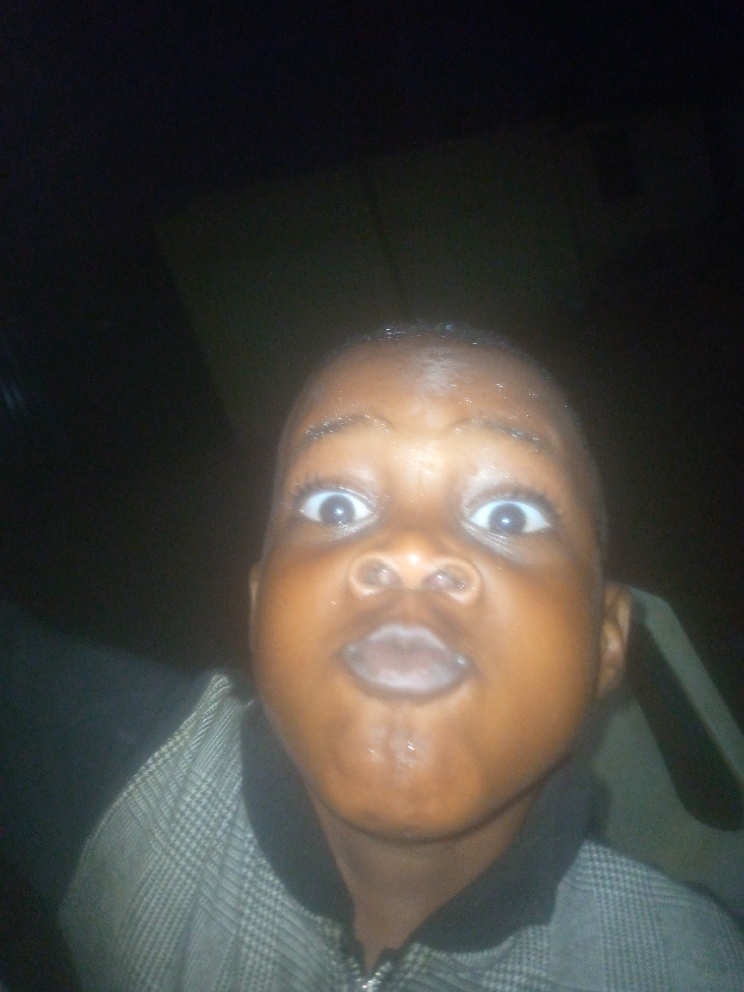 A close-up photograph shows a child with wide eyes and pursed lips. The child is wearing a patterned shirt with a dark collar. The background is dark and blurry. The location and city are unknown.