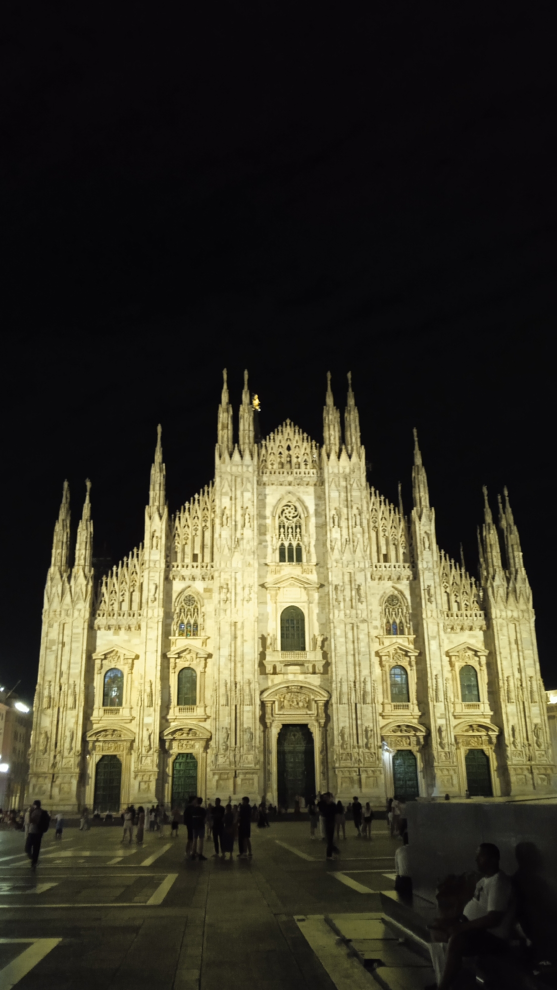 The image depicts the Milan Cathedral (Duomo di Milano) at night, illuminated by artificial lights. The cathedral is a grand Gothic structure with numerous spires and intricate architectural details. In the foreground, a paved piazza is visible, with several people walking and standing around. The sky above is dark and clear. The scene suggests a popular tourist area in Milan, Italy, where visitors gather to admire the illuminated cathedral after sunset.