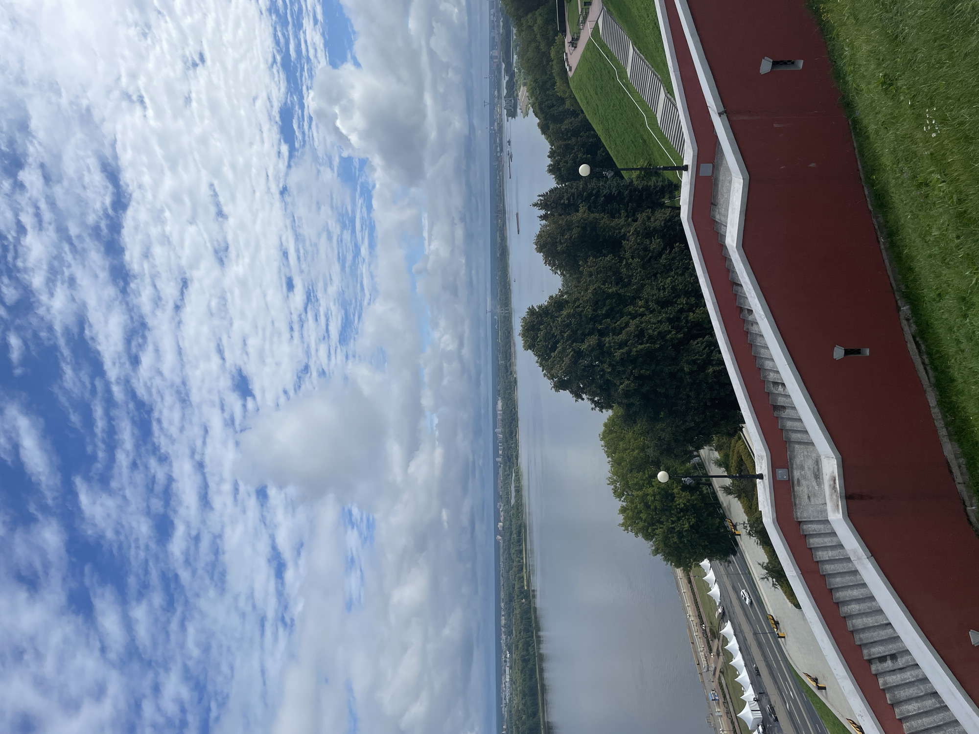 The image captures a view from a high vantage point, overlooking a wide river and the cityscape of Nizhny Novgorod, Russia. In the foreground, a prominent red-walled staircase with white trim dominates the lower half of the frame, leading down towards a road and the riverbank. The staircase is wide and appears to be made of concrete or stone, with numerous steps.
Beyond the staircase, a road runs parallel to the river, lined with white tents, yellow benches, and a few cars. The river itself is vast and calm, reflecting the cloudy sky. Several barges and boats are visible on the water. On the opposite bank of the river, the city skyline is visible, characterized by a mix of buildings and trees. The distant horizon is dotted with industrial structures and possibly bridges.
The sky is a mix of blue and white, with fluffy clouds scattered across it, suggesting a partly cloudy day. Lush green grass covers the embankment to the right of the staircase, with some scattered wildflowers. Trees, primarily deciduous and dark green, are visible on the slope leading down to the river and also on the opposite bank. The overall impression is a scenic, urban landscape with a strong emphasis on the river and the architectural elements of the viewpoint.