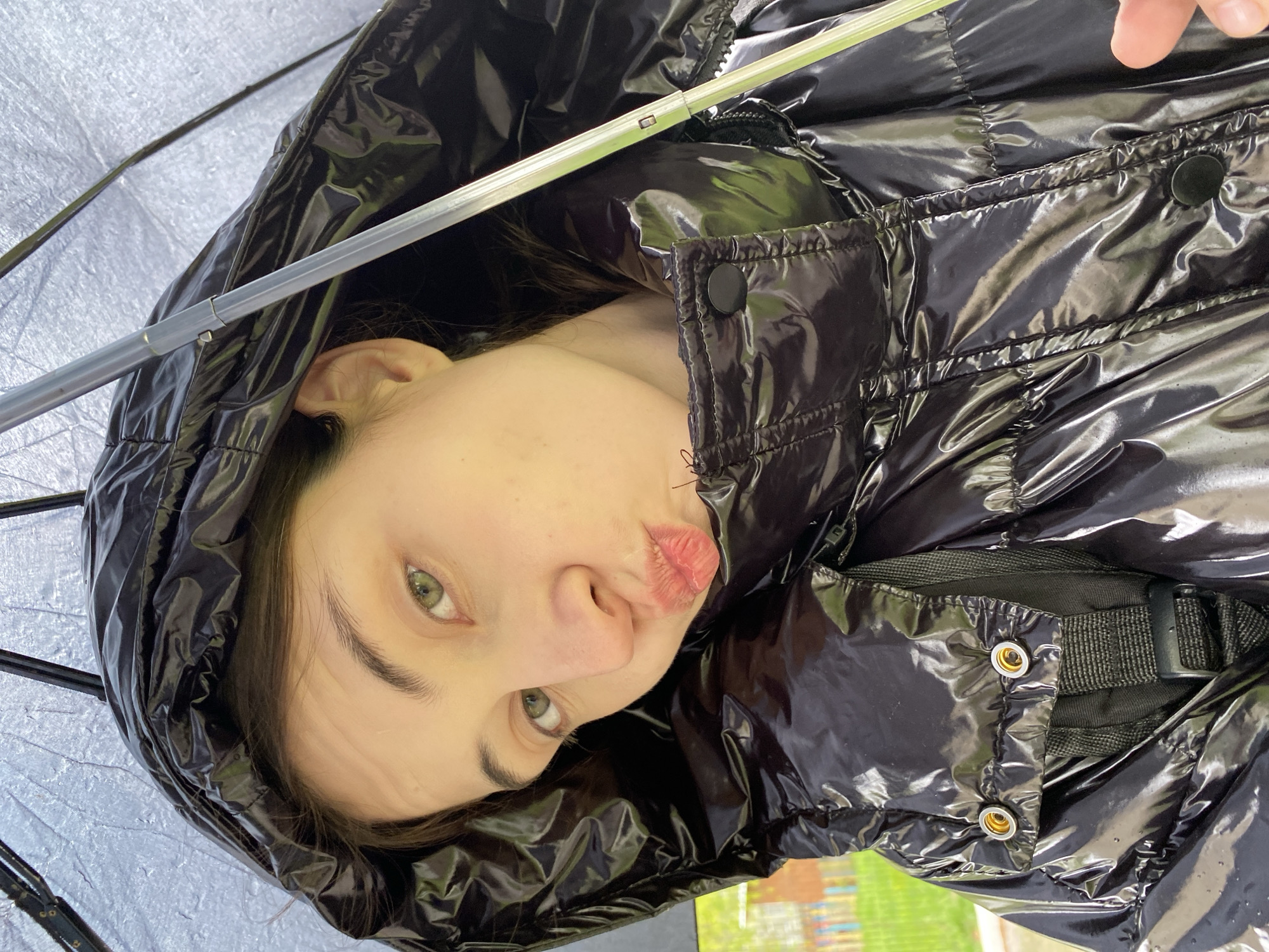 A young person with fair skin, green eyes, and pursed lips is depicted in this close-up, low-angle shot. The individual is wearing a shiny, black, hooded puffer jacket. The jacket's hood is pulled up, partially obscuring the person's hair. The environment suggests an outdoor setting under a translucent umbrella, given the presence of the umbrella's ribs and canopy visible in the frame. The background shows blurred elements that suggest a natural outdoor scene, possibly with greenery and some structures. This image does not contain enough information to determine the specific location in the city of Votkinsk, Russia.