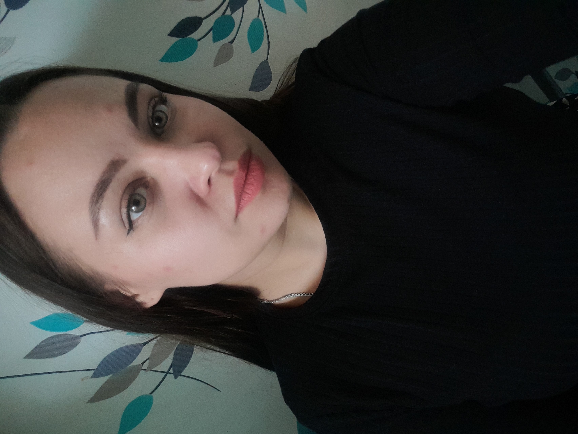 A close-up selfie of a young woman with fair skin, brown hair, and green eyes. She has noticeable dark eyeliner and light pink lipstick. The woman is wearing a black, ribbed top and a thin silver necklace. The background is a light-colored wall with a repeating leaf pattern in shades of teal, blue, and gray. The location and city are unknown.