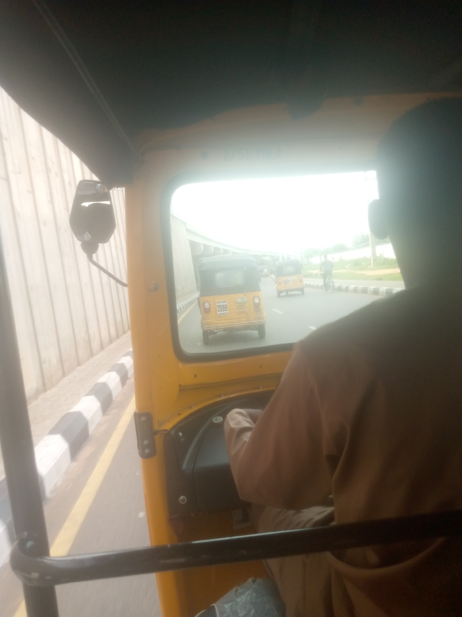 The image is a first-person perspective from inside a yellow auto-rickshaw, likely in Wauro Jabbe, Nigeria, given the context. The driver, a person wearing a brown garment, is partially visible in the foreground, operating the vehicle. Outside the auto-rickshaw's windshield, the road is visible, with another yellow auto-rickshaw in front, displaying the license plate "7099". Further down the road, a person on a bicycle is also visible. The surrounding environment includes a light-colored wall or structure on the left, and a pavement with black and white striped borders. The sky appears bright and possibly overcast, contributing to the overall washed-out lighting of the scene.