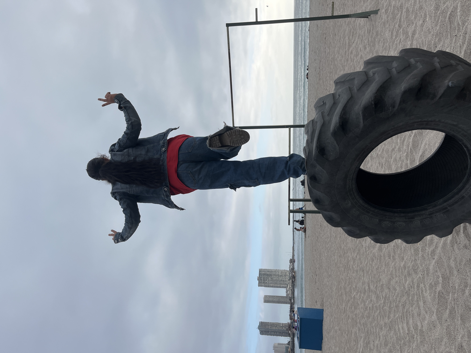 In Iquique, Chile, on a sandy beach, a person stands on a large tire, balancing on a horizontal bar. They are wearing a denim jacket, a red shirt, and ripped blue jeans, with their right leg extended. Their arms are raised, with two fingers on each hand extended in a peace sign. The background shows the ocean, a few buildings, and other people on the beach.