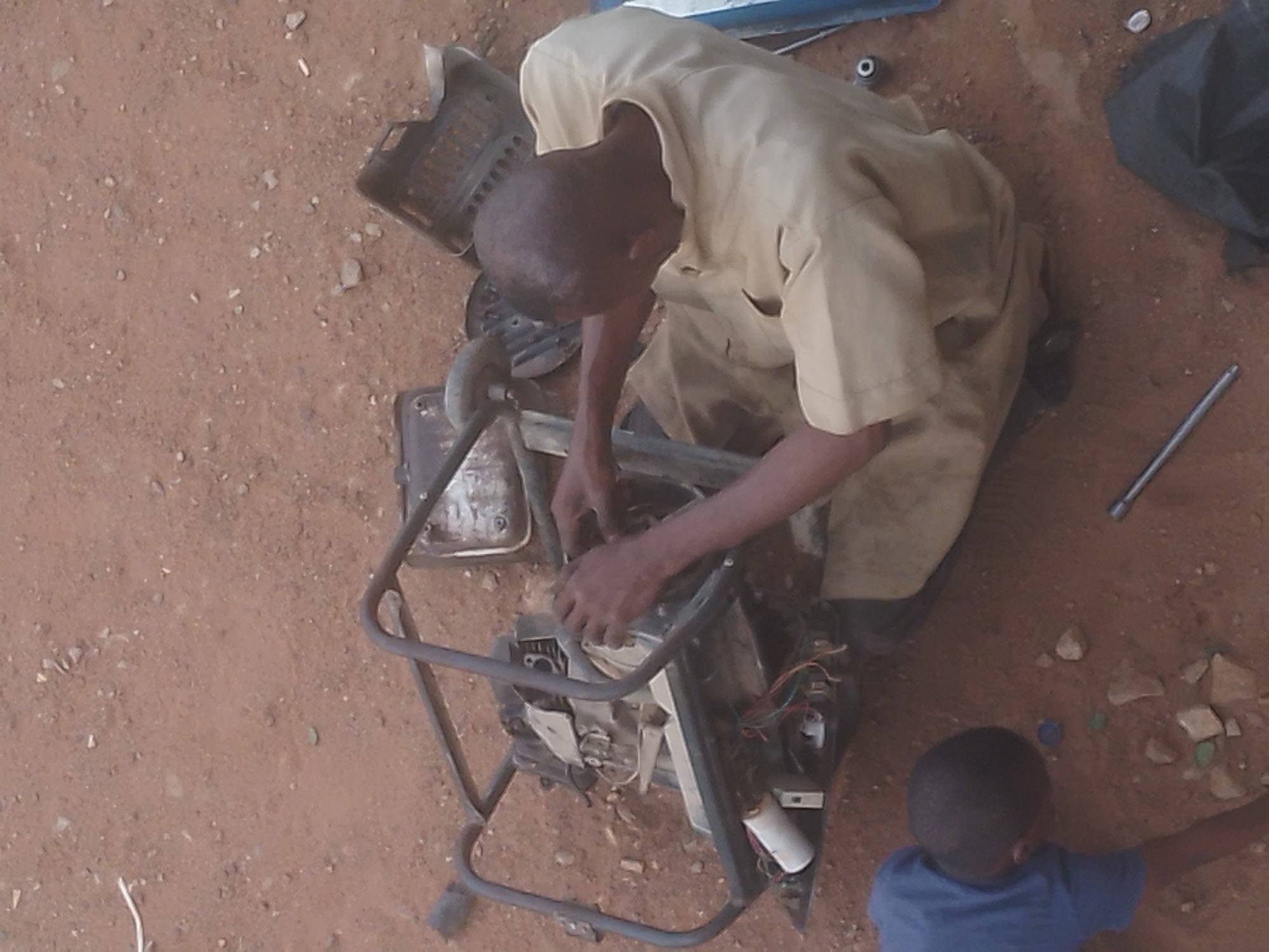 The image shows a person wearing a tan or light-colored shirt repairing a piece of machinery, possibly a generator, in an outdoor environment with reddish-brown dirt or soil visible. A child in a blue shirt is also present, partially visible in the lower right corner of the frame. Miscellaneous parts, tools, and debris are scattered around the area. The location is Gura Top, Nigeria.