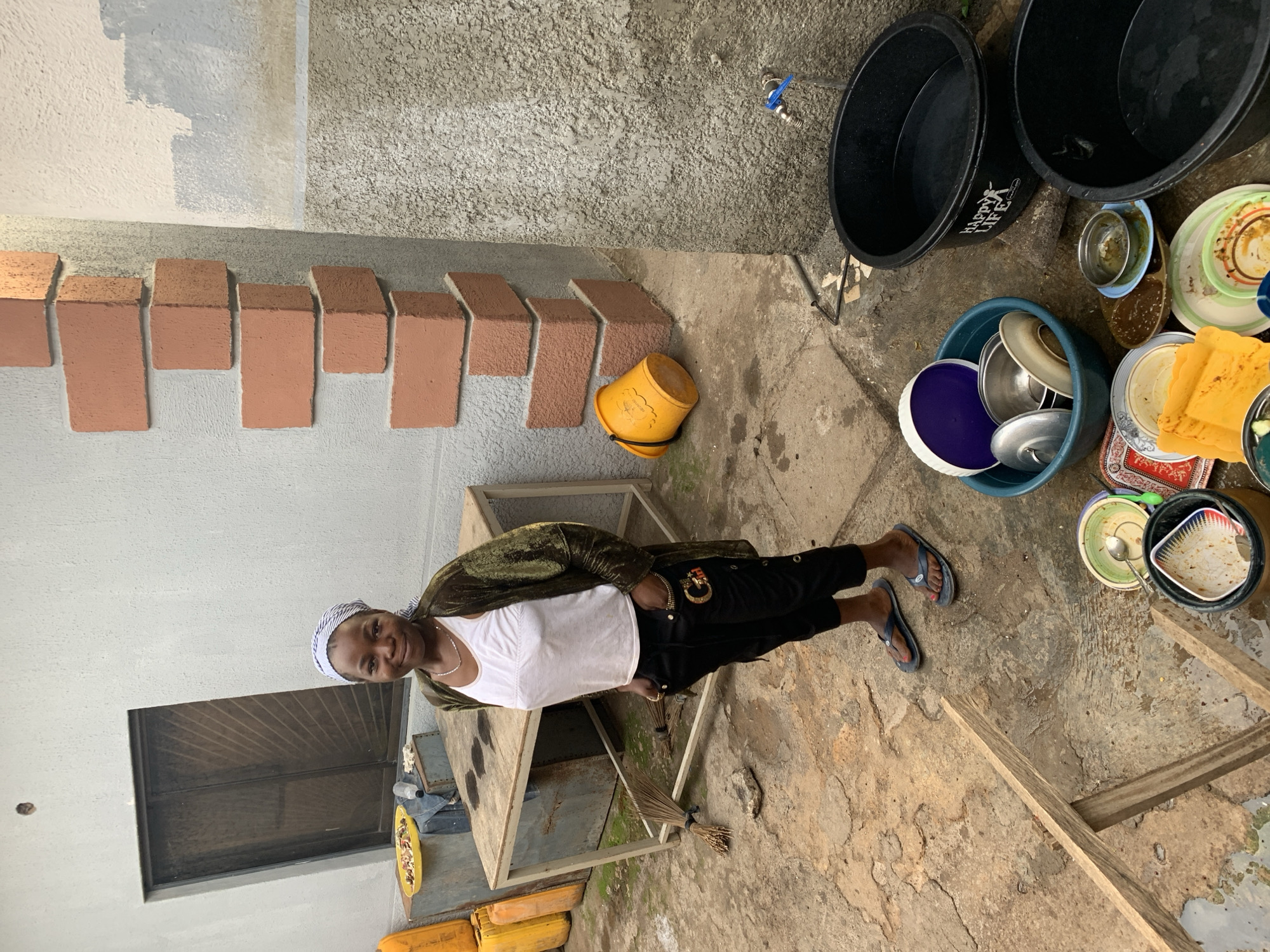 A woman stands in a courtyard with concrete walls, one of which is decorated with rows of protruding rectangular bricks. She wears a white top, black pants, and a green jacket. She has a headscarf. The courtyard contains household items including black buckets, a blue basin filled with dishes, and plates. There's a yellow bucket near the wall and a wooden table is in the foreground. The scene appears to be in a residential area, potentially in Jos, Nigeria. The ground is concrete with visible cracks and stains.