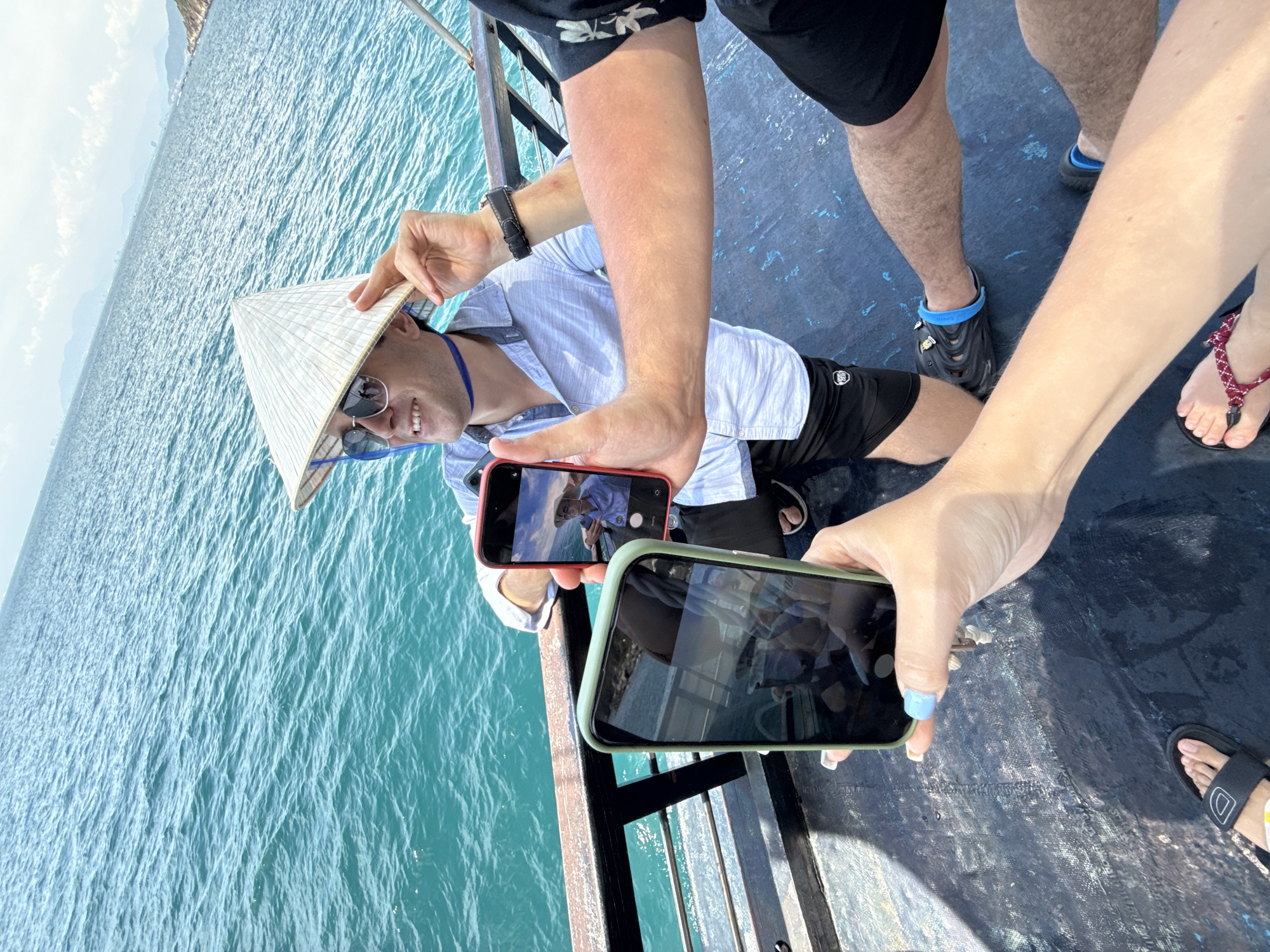 Captured from an elevated perspective on a vessel, the image presents a scene where a man lies prone on the deck, sporting a conical Asian hat and sunglasses. This individual is the focus of attention as at least two people use their mobile phones to photograph him. The setting appears to be a boat ride on a sunny day, with an expanse of rippling turquoise water visible in the background. The location is identified as being in the city of Bac Nha Trang Ward, Vietnam. The image also includes portions of other people, including bare legs, arms, and feet, suggesting a shared experience or tour group setting.