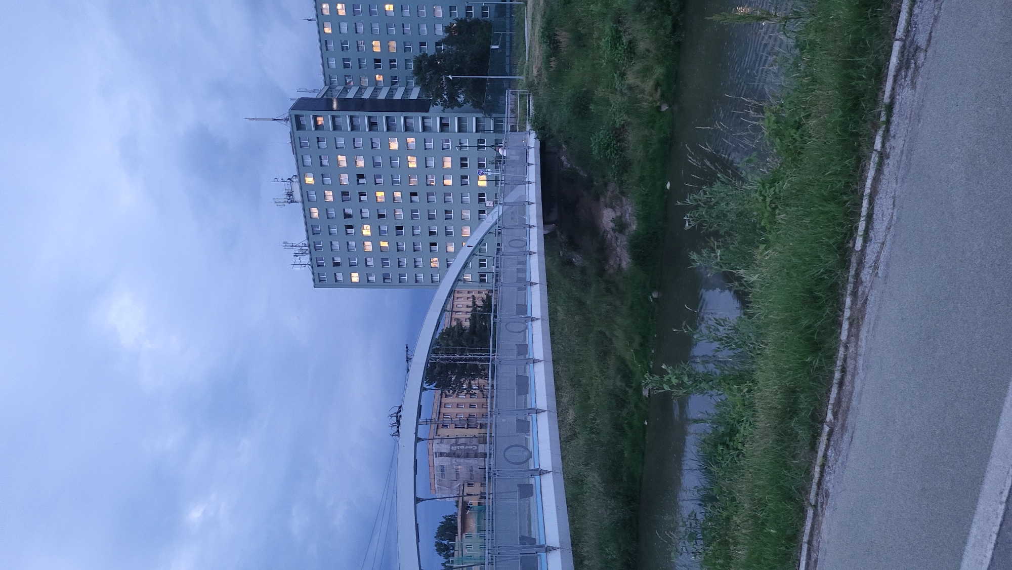 The image shows a wide, modern pedestrian bridge arching over a narrow river or canal in the city of Prešov, Slovakia. The bridge has a white railing with vertical supports and a blue stripe along its length. The river is lined with dense green grass and some bushes. In the background, several tall, light gray apartment buildings are visible, their windows illuminated, suggesting it is dusk or evening. The sky is overcast with a deep blue and gray tone. The apartment buildings appear to be part of a residential complex, possibly located in a suburban area of Prešov. The scene is tranquil and urban.