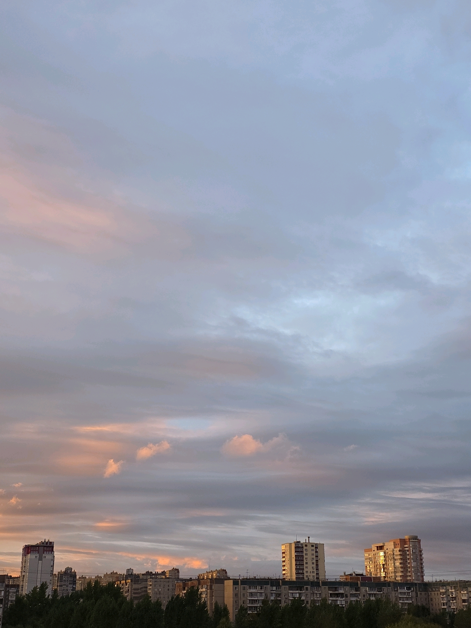 The image depicts a skyline with apartment buildings and a vast sky filled with clouds tinged with the colors of sunset. The buildings are varied in height and architectural style, with some featuring balconies and numerous windows. A line of trees runs along the bottom of the image, separating the buildings from the foreground. The sky is a mix of soft blues, grays, and warm pink and orange hues from the setting sun, creating a serene and atmospheric scene. Without specific markers or landmarks visible, the exact location within Chelyabinsk, Russia, cannot be determined from the image alone. The image captures a quiet moment as day transitions into evening in an urban environment.