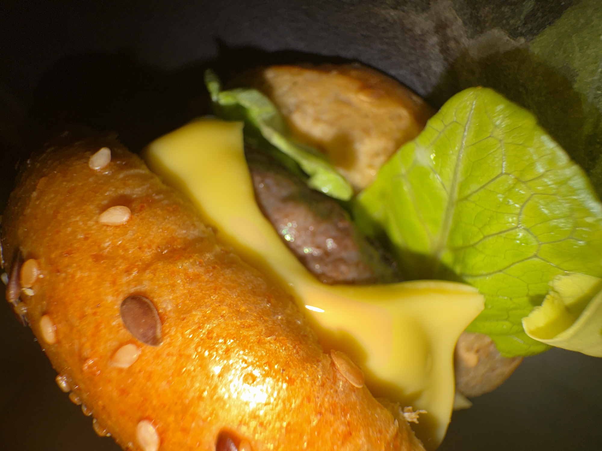 The image depicts a close-up shot of a burger. The top bun is visible, featuring sesame and other seeds. A slice of yellow cheese is partially melted over the burger patty. Lettuce leaves are also present, adding a green element to the image. The other components of the burger are slightly obscured. The image is taken indoors, possibly in a restaurant or at home, but there's no specific indication of the location being in Sofia, Bulgaria.